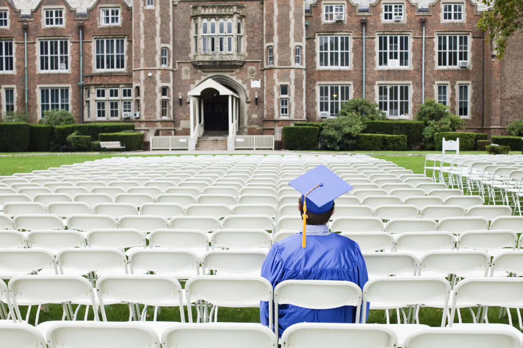 A graduate in a blue cap and gown sits among empty white chairs, facing a historic brick building, ready for the commencement ceremony.