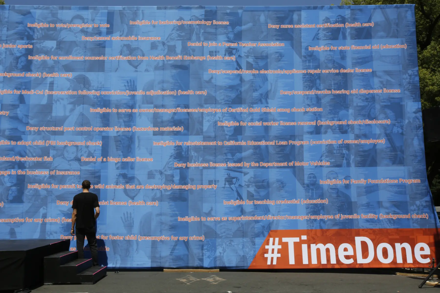 A person stands in front of a large blue backdrop covered with text listing various disqualifications and barriers, labeled #TimeDone.