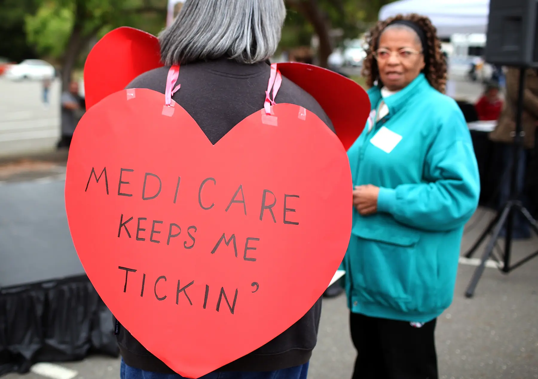 A person wearing a large red heart-shaped sign reads "Medicare keeps me tickin’," while another individual in a turquoise jacket looks on.