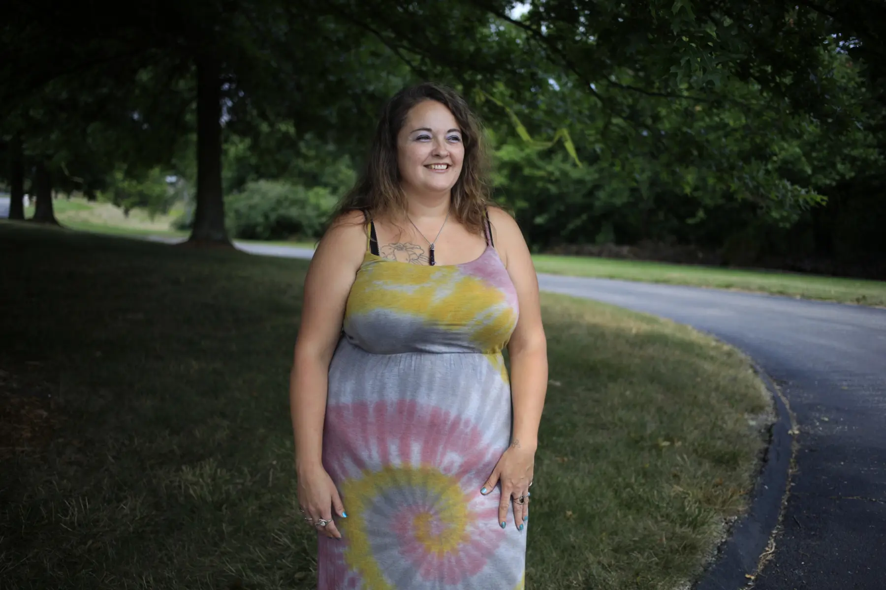 Smiling woman stands outdoors in a colorful tie-dye dress, surrounded by lush greenery and a winding path in the background.