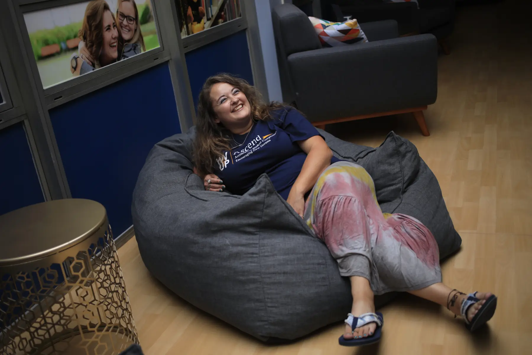 A woman with long, wavy hair reclines in a large gray beanbag chair, wearing a blue shirt and colorful pants, smiling joyfully.