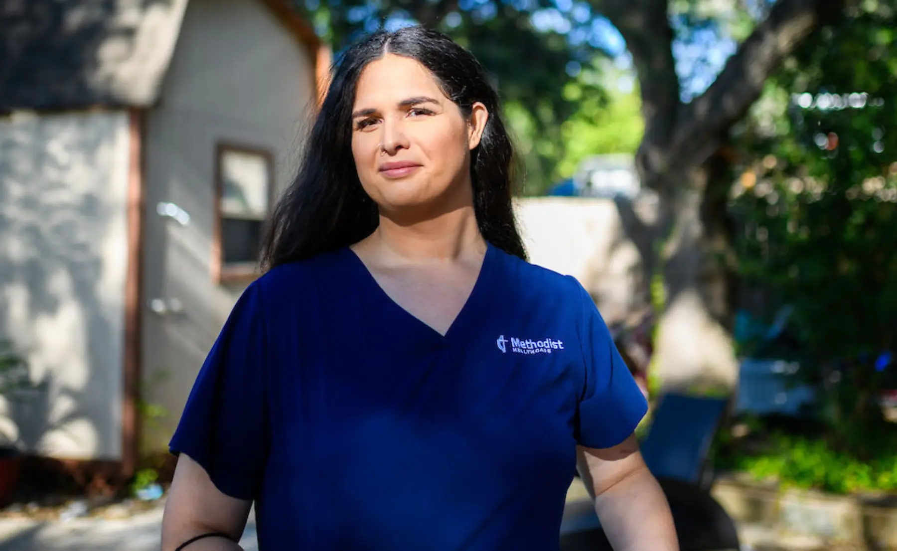 A woman with long dark hair smiles confidently while wearing a blue scrubs top in a bright outdoor setting with greenery.