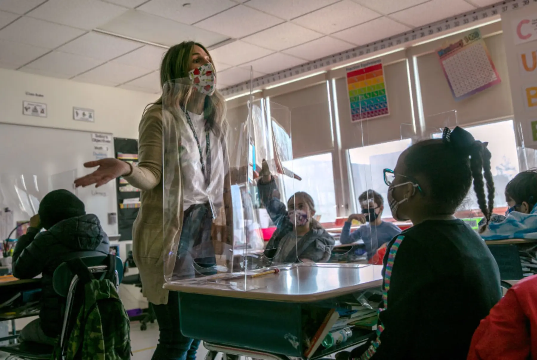 Teacher wearing mask stands at front of classroom with students sitting at desks.