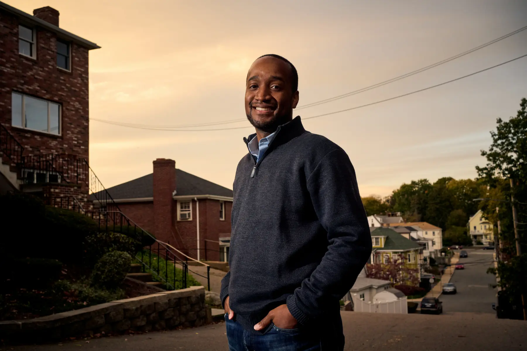 A smiling man stands confidently on a sloped street, dressed in a dark zip-up sweater, with houses and trees in the background.