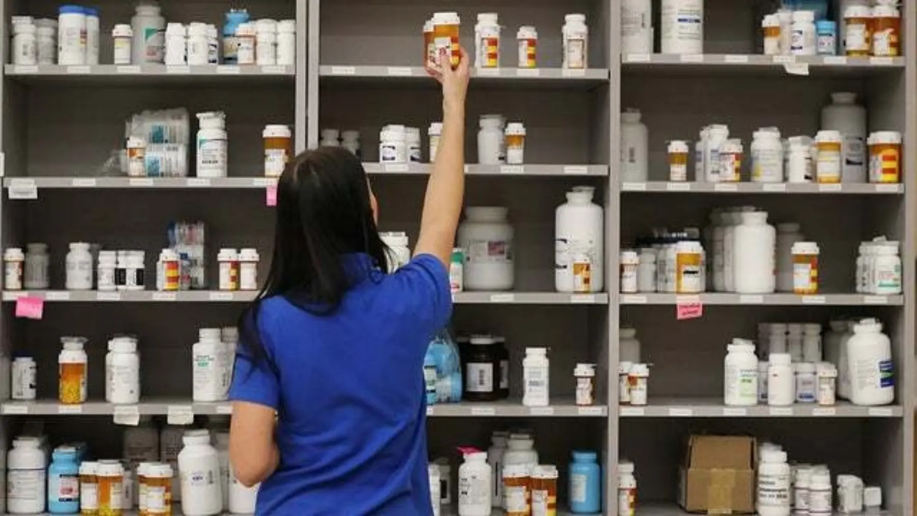A pharmacy employee in a blue shirt reaches for a prescription bottle on a shelf filled with various labeled medicine containers.
