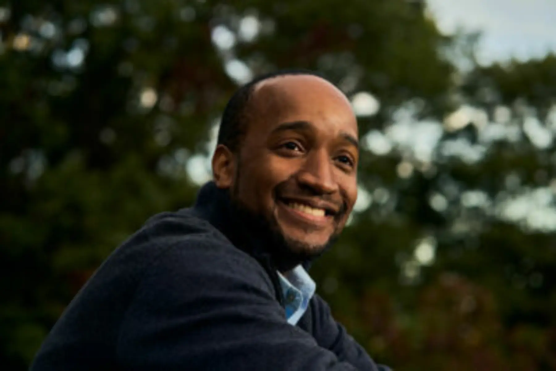 A man with a warm smile sits outdoors, wearing a dark sweater, with trees blurred in the background, capturing a serene moment.