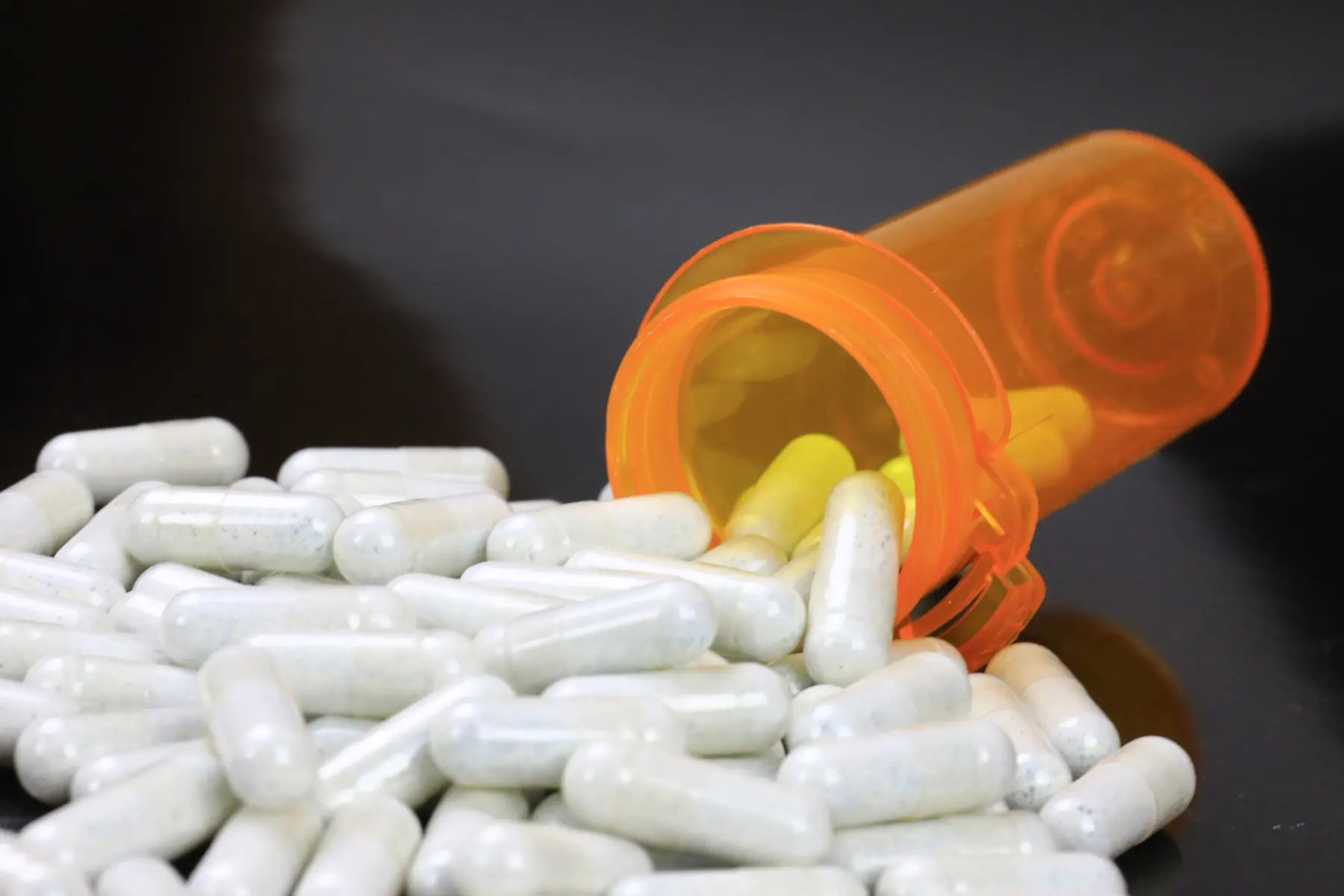 Capsules spill from an orange prescription bottle onto a reflective black surface, creating a disordered yet visually striking scene.