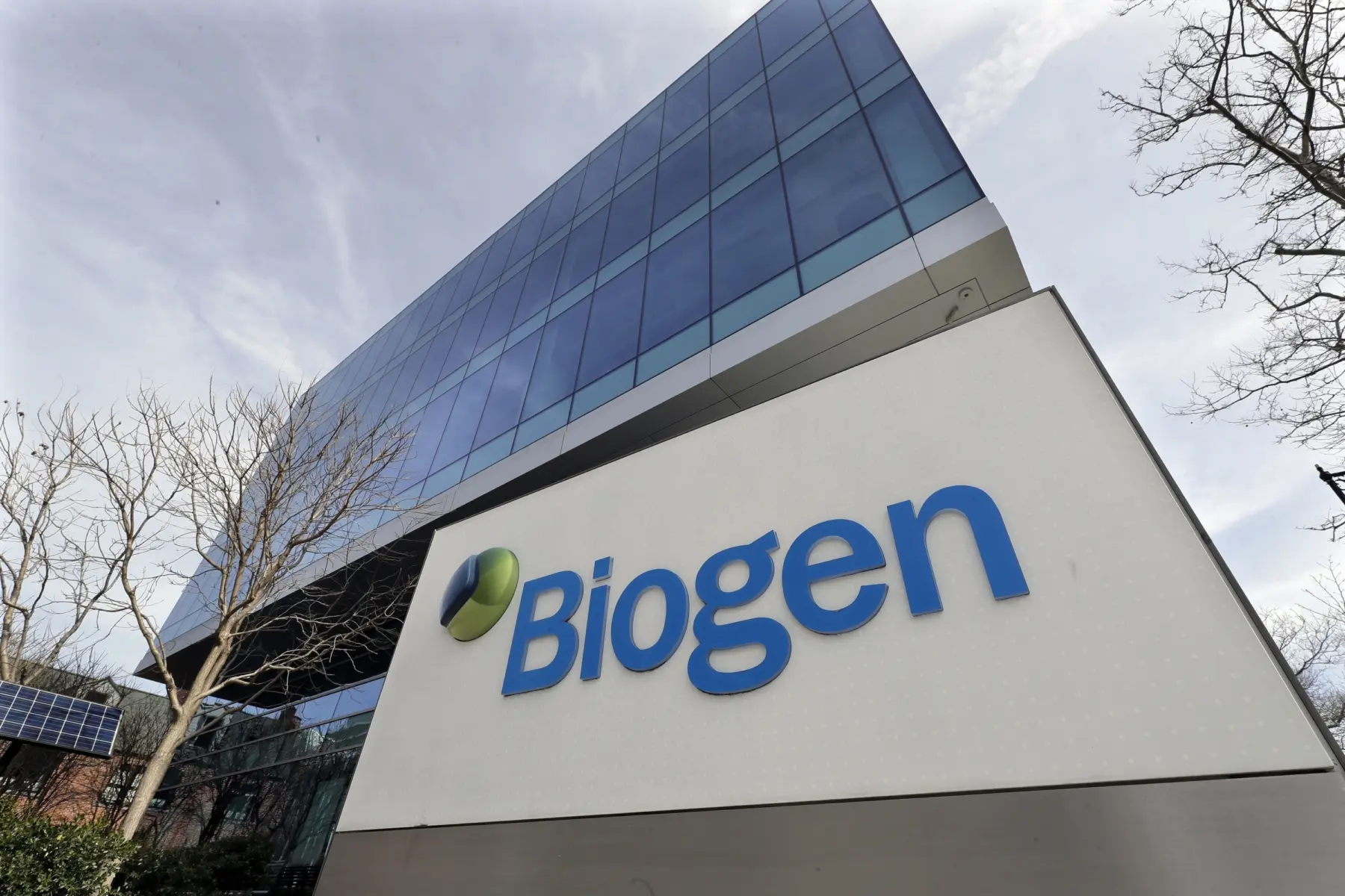 A modern glass building with a prominent Biogen sign in blue, surrounded by bare trees and solar panels, under a cloudy sky.
