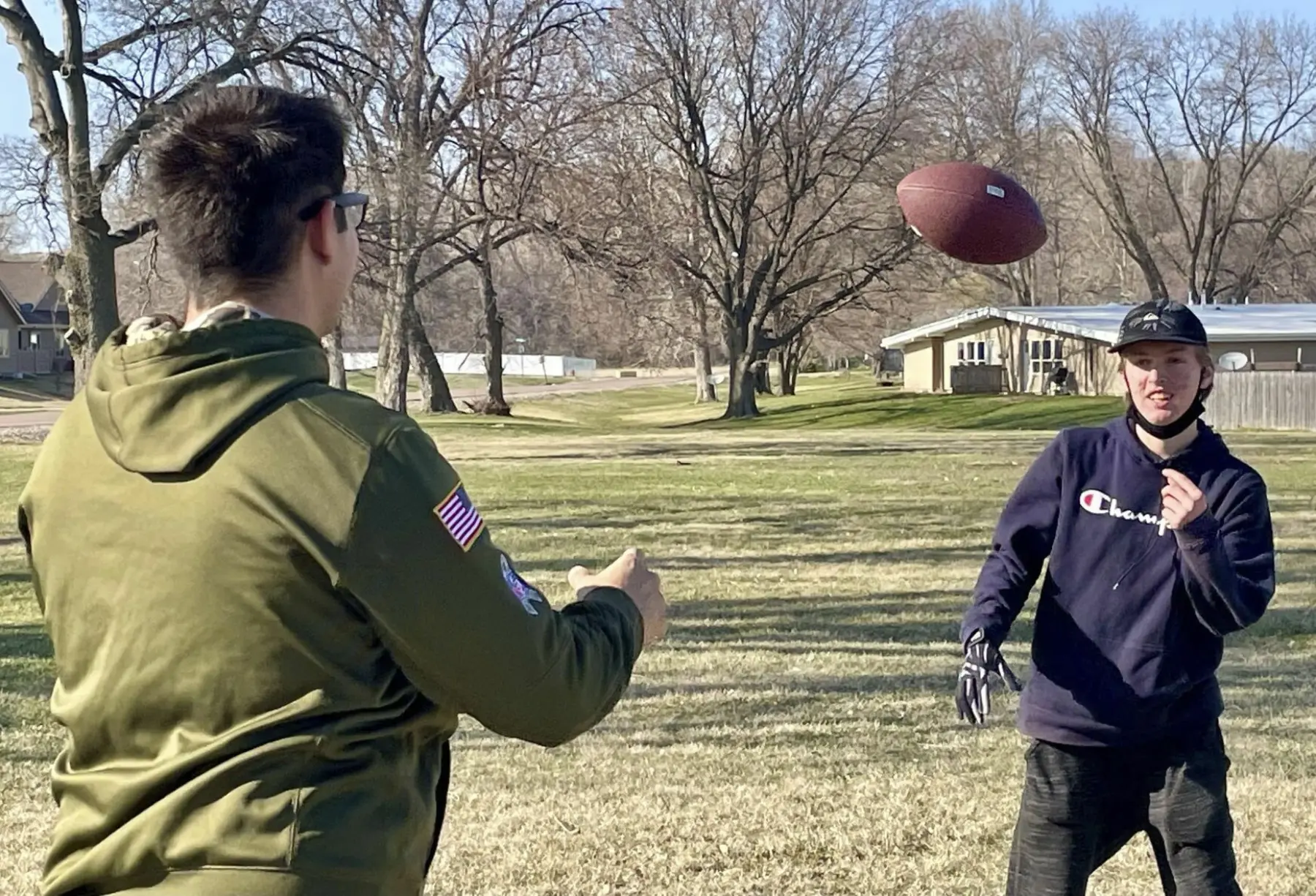 Two young men play catch with a football in a grassy field, surrounded by bare trees and suburban homes in the background.