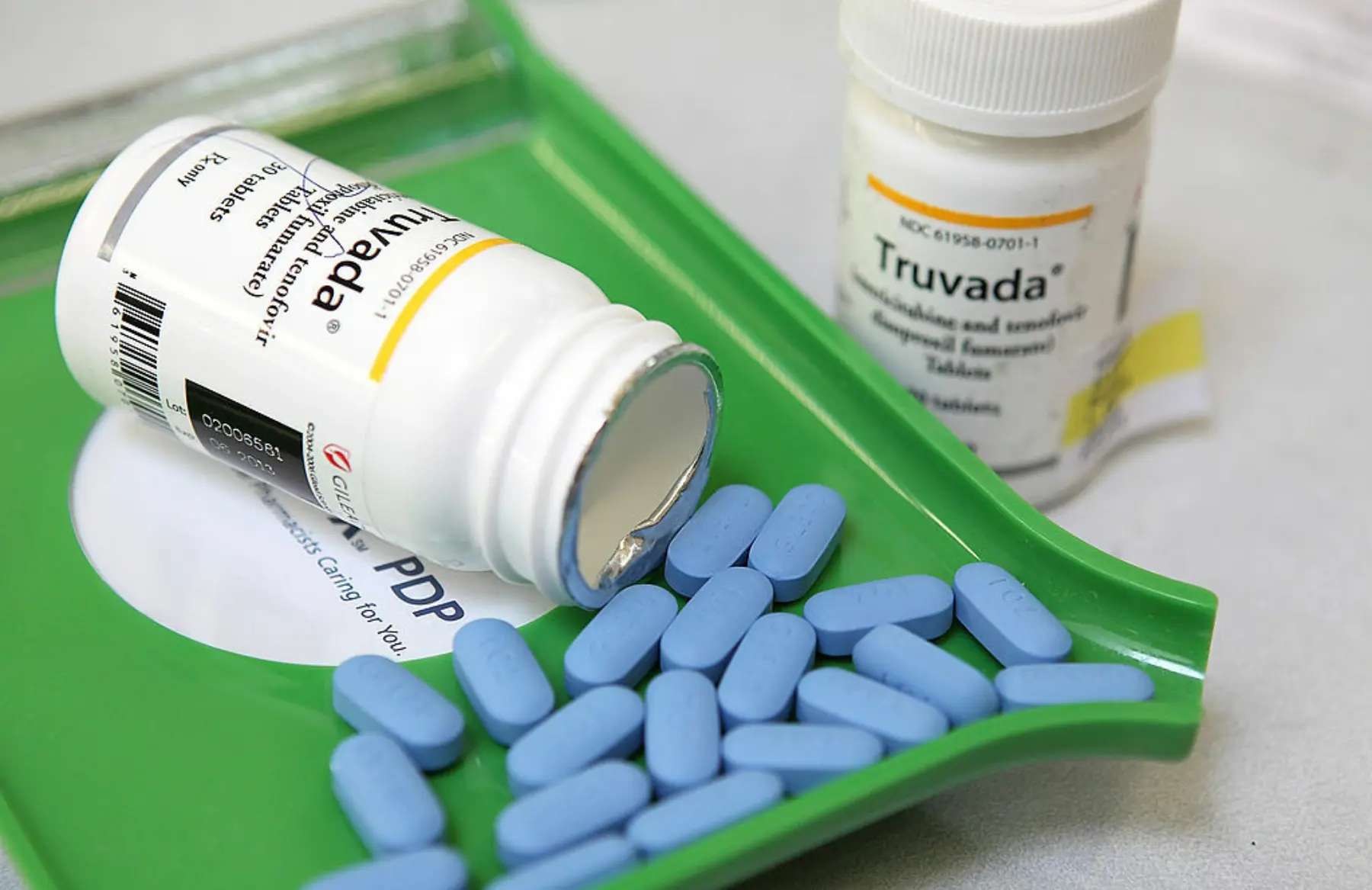 A green pill tray holds several blue oval pills labeled Truvada, alongside a nearby white prescription bottle.