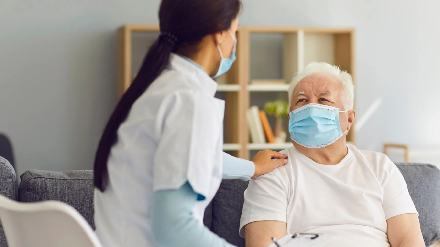A healthcare professional in a mask engages with an elderly patient, both wearing masks, in a comfortable, well-lit room.