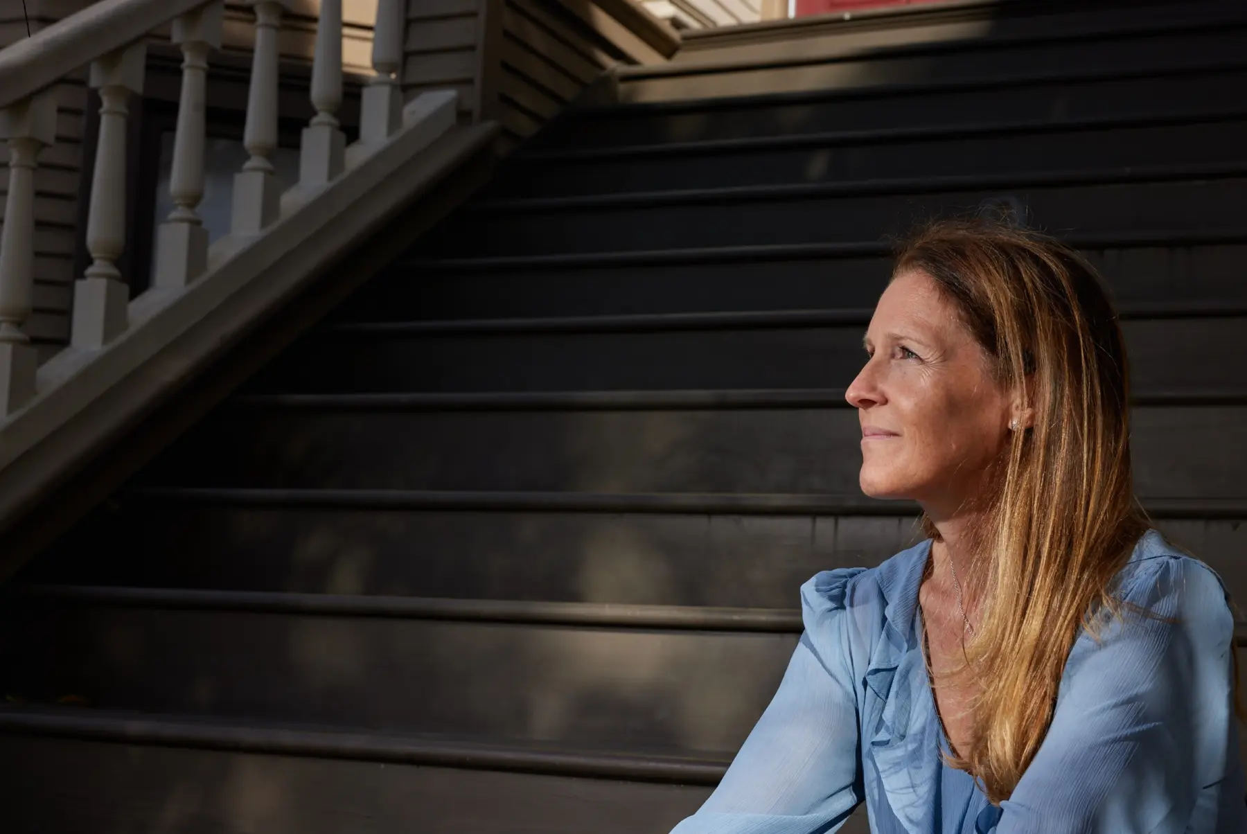 A woman with long, brown hair sits on black wooden stairs, gazing thoughtfully into the distance, dressed in a light blue blouse.