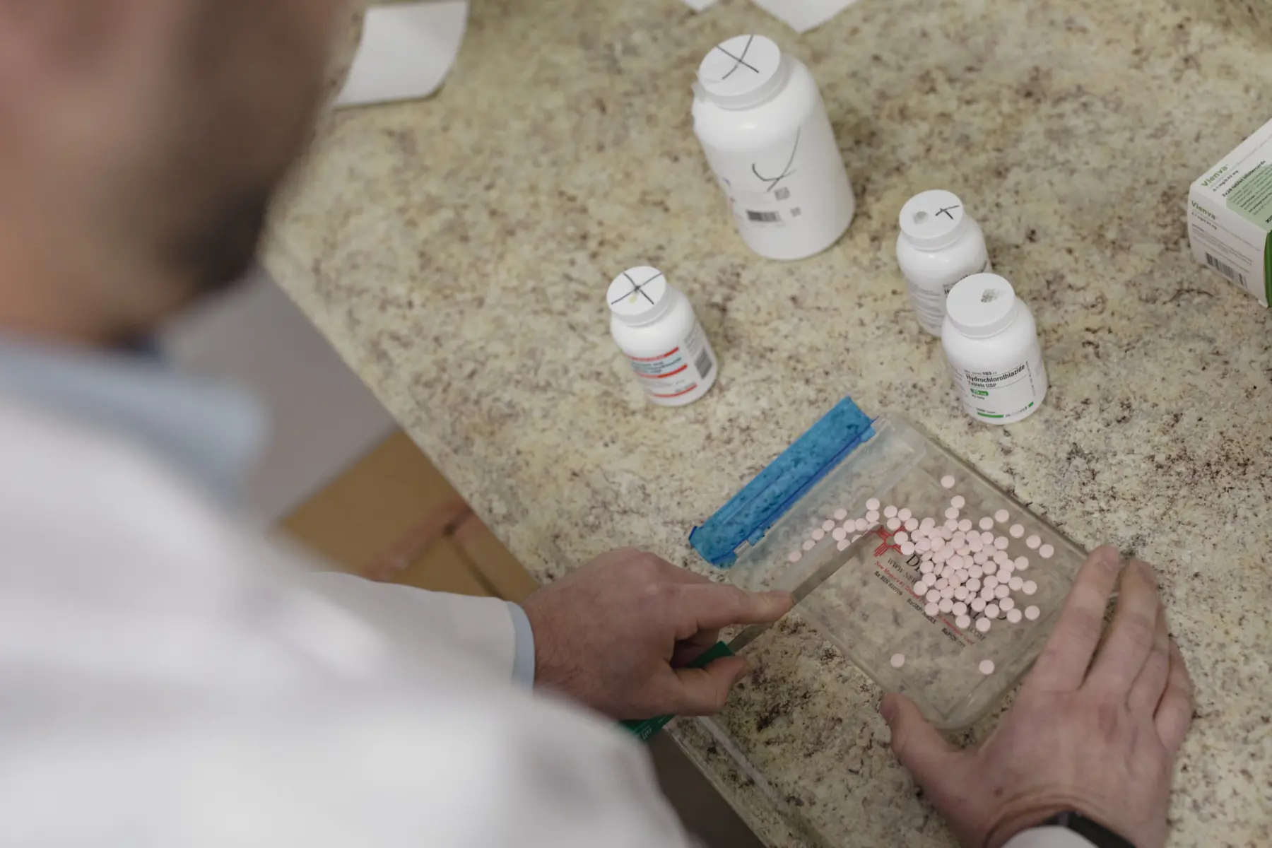 A healthcare professional in a lab coat is counting pink tablets on a tray, surrounded by various bottles of medication on a countertop.