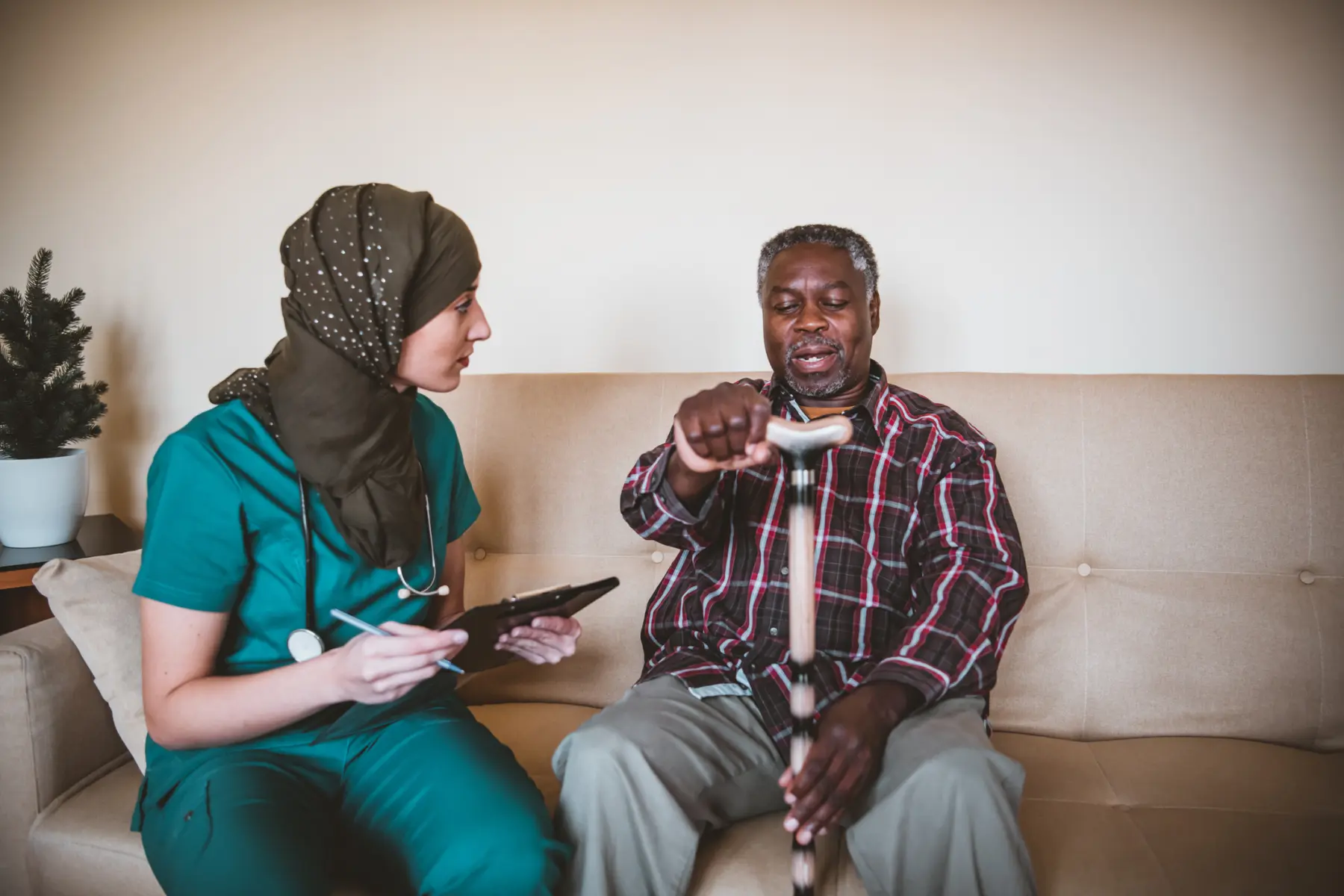 Woman in scrubs sits on couch with a patient holding a cane.