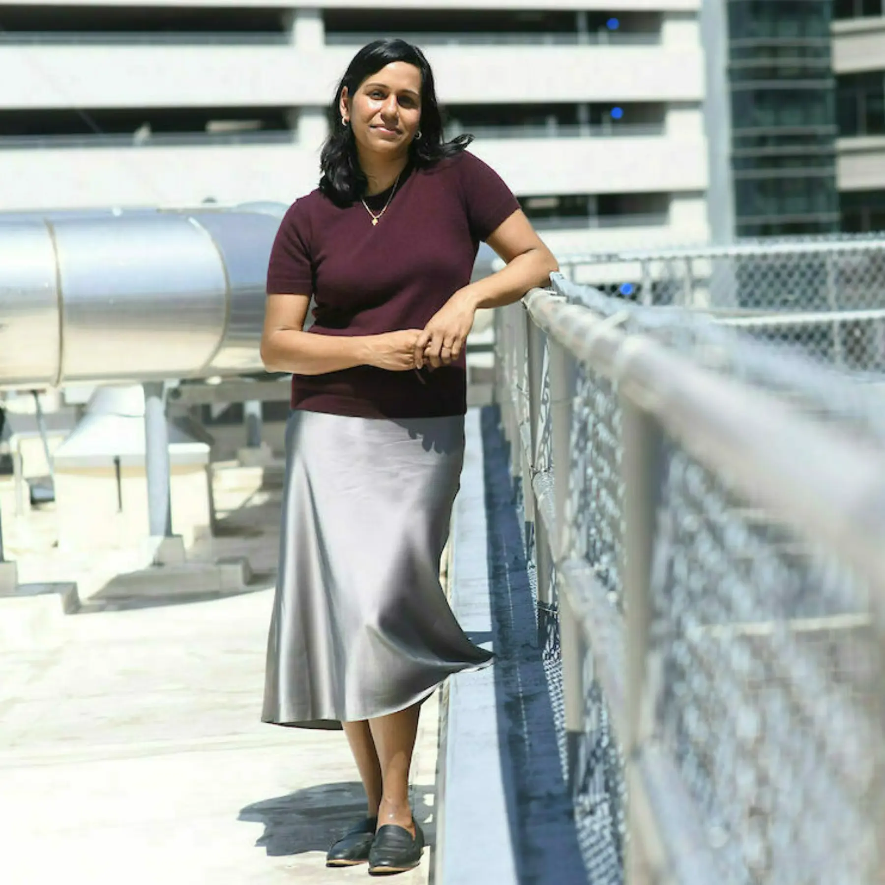 Woman stands on rooftop of building leaning on chain link fence.