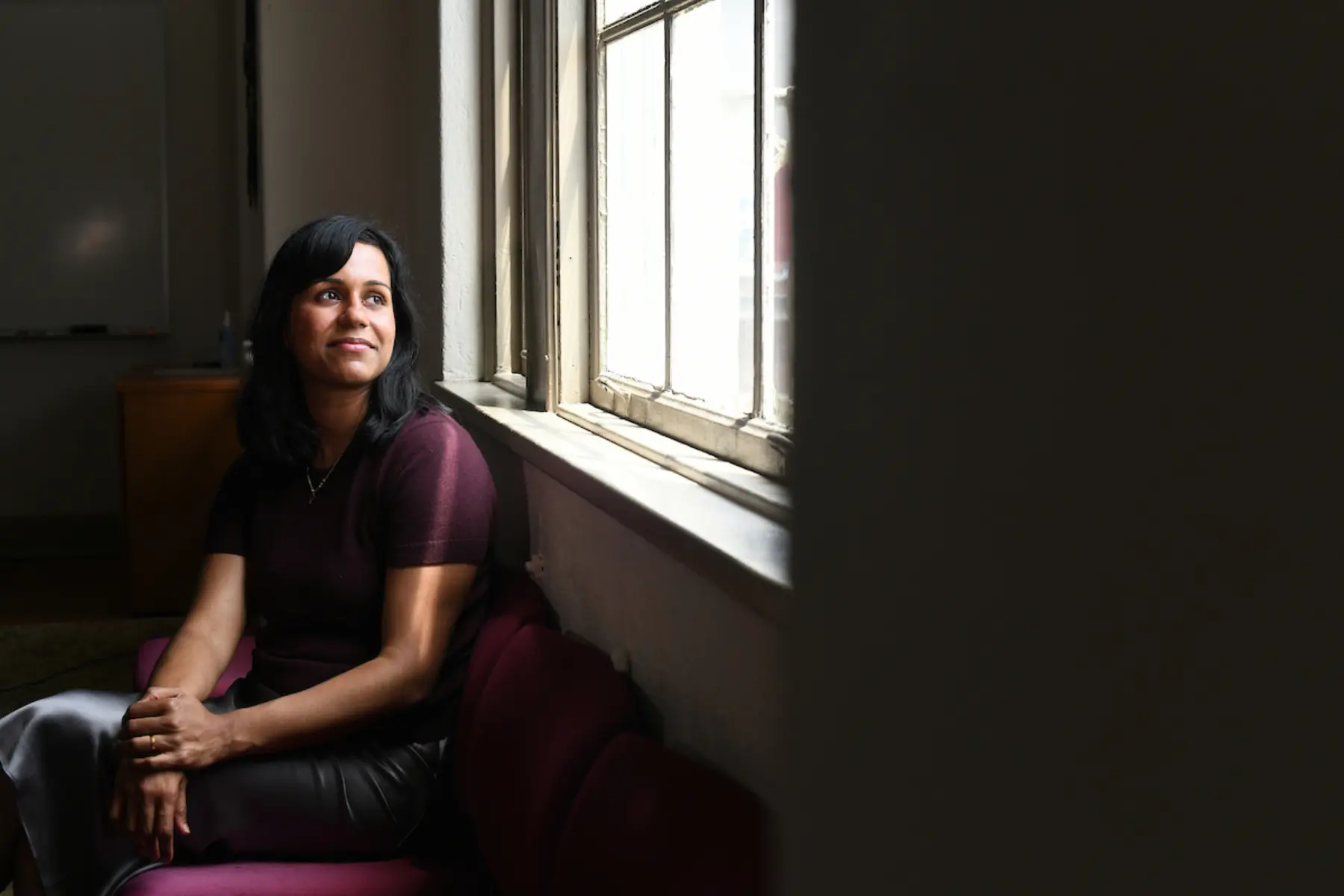 Woman sits looking out a bright window as rest of room is dark.