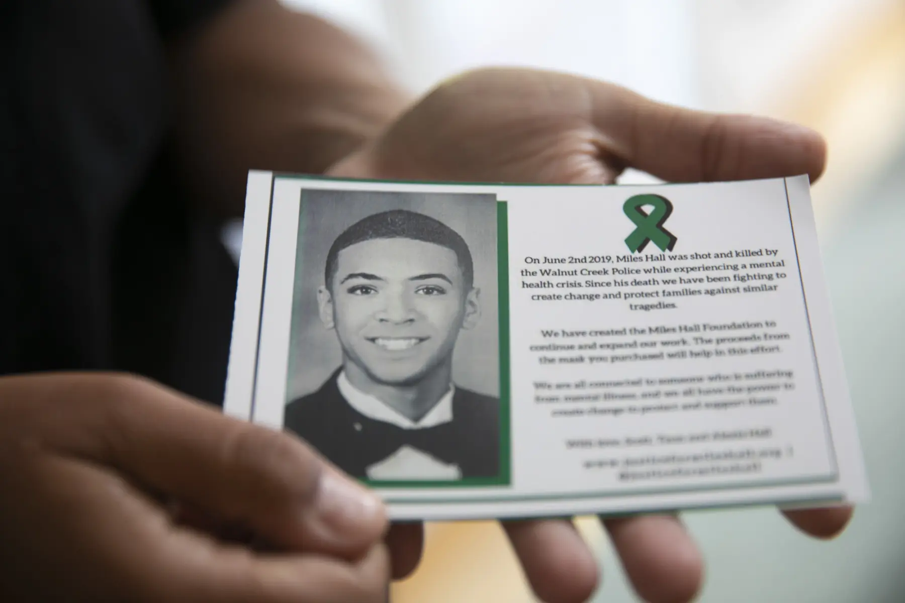 A hand holds a card honoring Miles Hall, featuring his photo, a green ribbon, and text about addressing mental health issues after his tragedy.