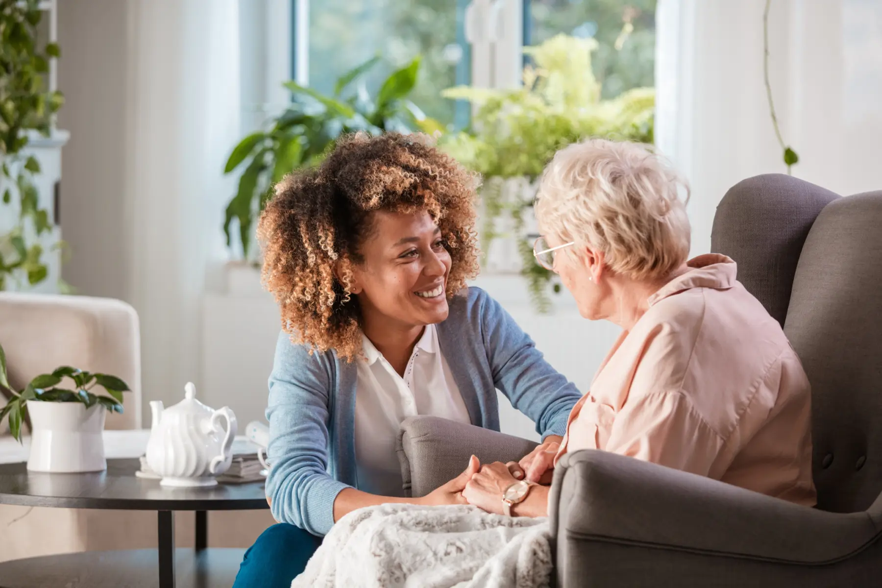 A young woman with curly hair smiles warmly while holding the hands of an elderly woman in a cozy, plant-filled room.