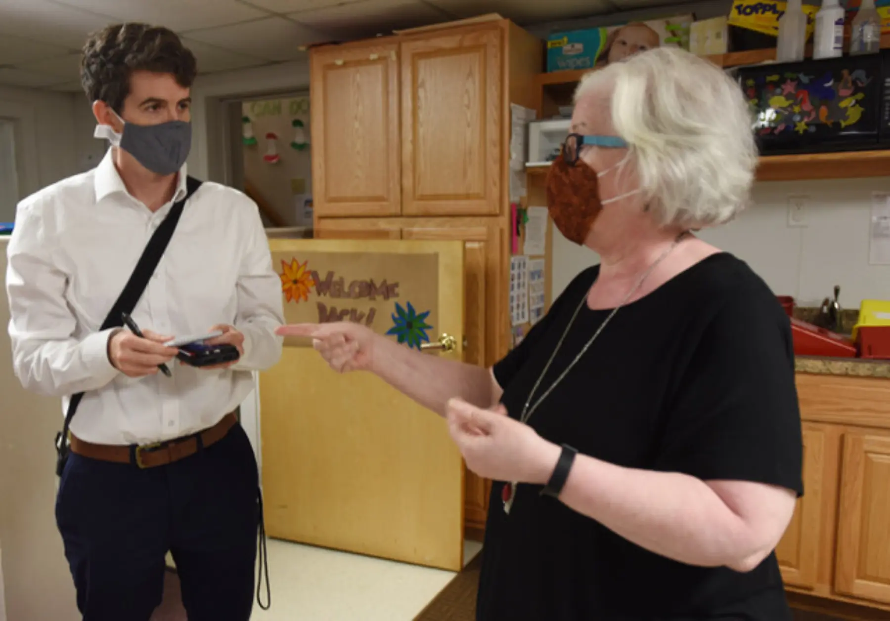 A reporter with a notebook and a face mask stands in the kitchen takes notes while interviewing a woman who gestures with her hands.