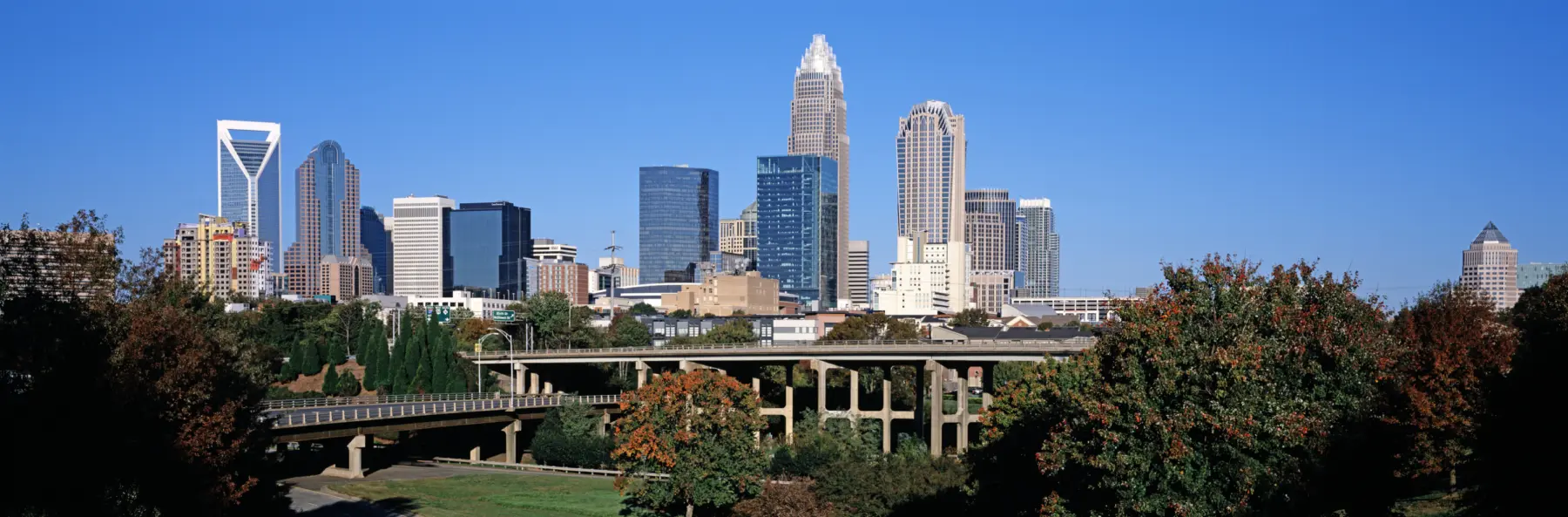 Skyline of Charlotte, North Carolina shows skyscrapers, trees and highways against a bright blue sky.