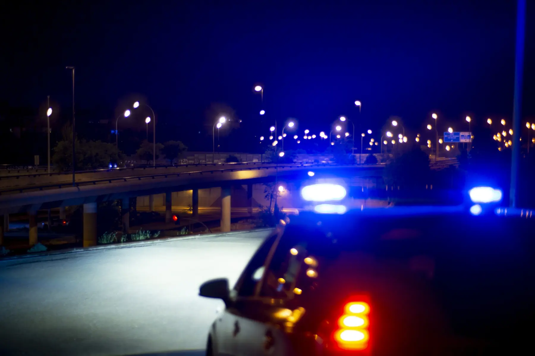 Emergency lights illuminate a dark urban highway at night, creating a vibrant contrast against the calm surroundings and distant streetlights.