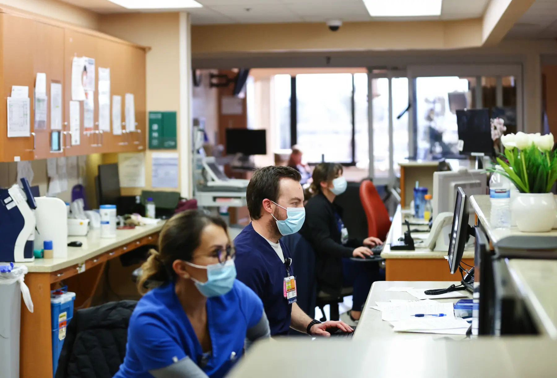 Healthcare professionals in scrubs and masks work at computers in a busy hospital environment, with documents and plants on the counter.
