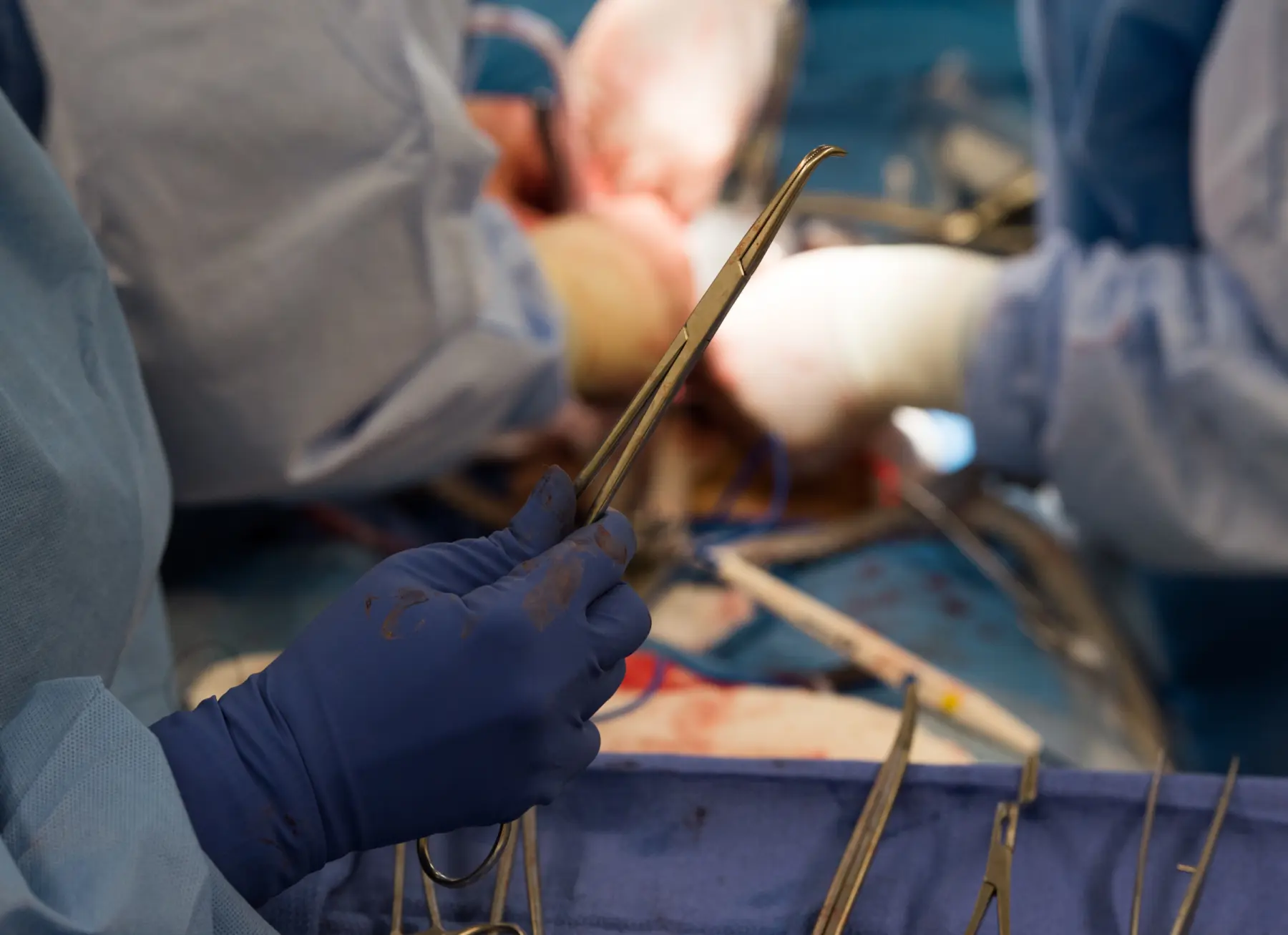 Hands in blue surgical gloves hold a surgical instrument, surrounded by various tools and a blurred surgical field in the background.