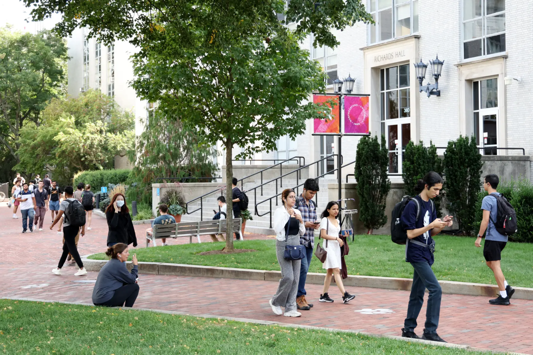 Students walk along a brick path in a university setting, with greenery and modern buildings in the background, engaged in various activities.