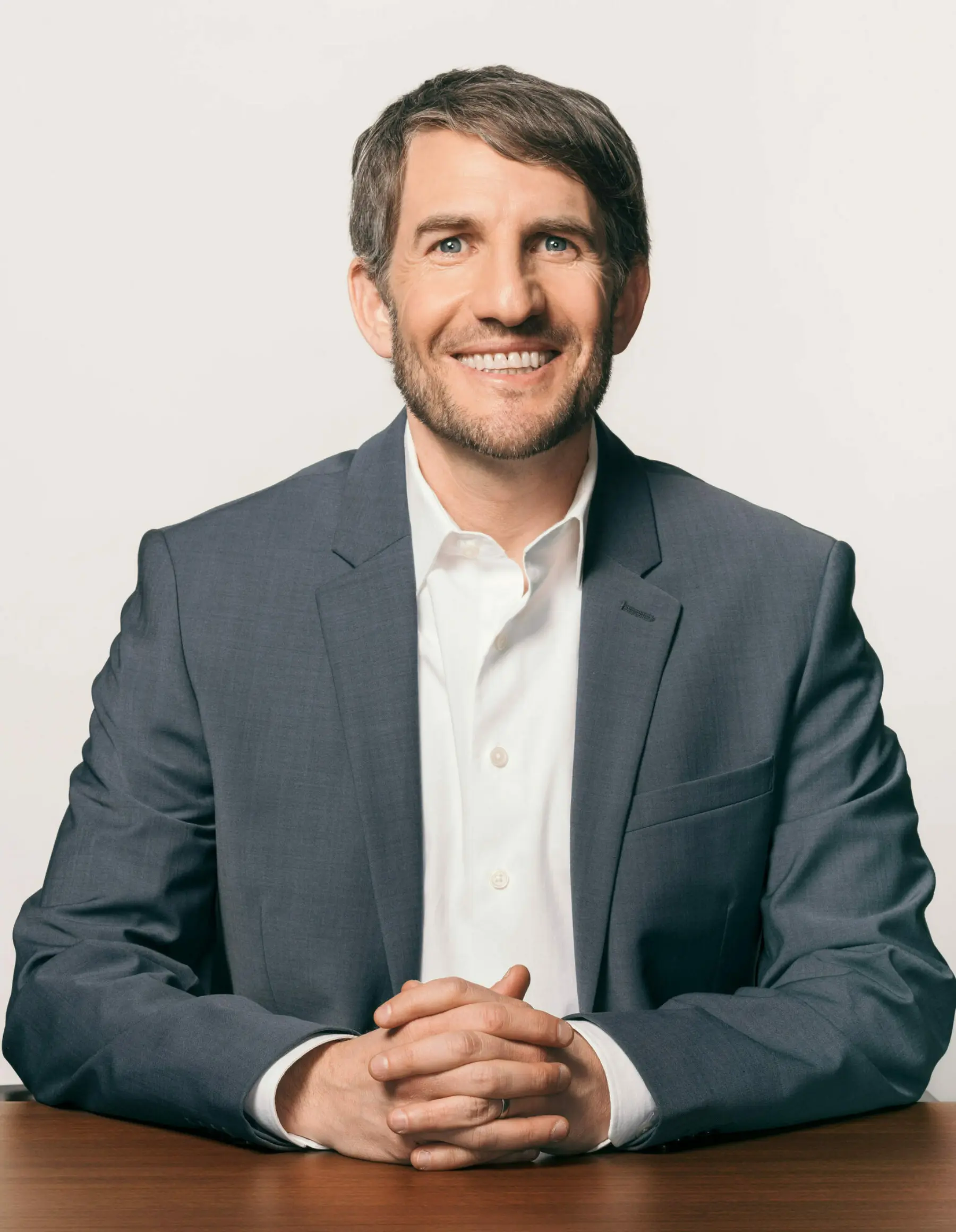 Smiling man in a gray suit and white shirt sits at a wooden table with hands clasped, against a light background.