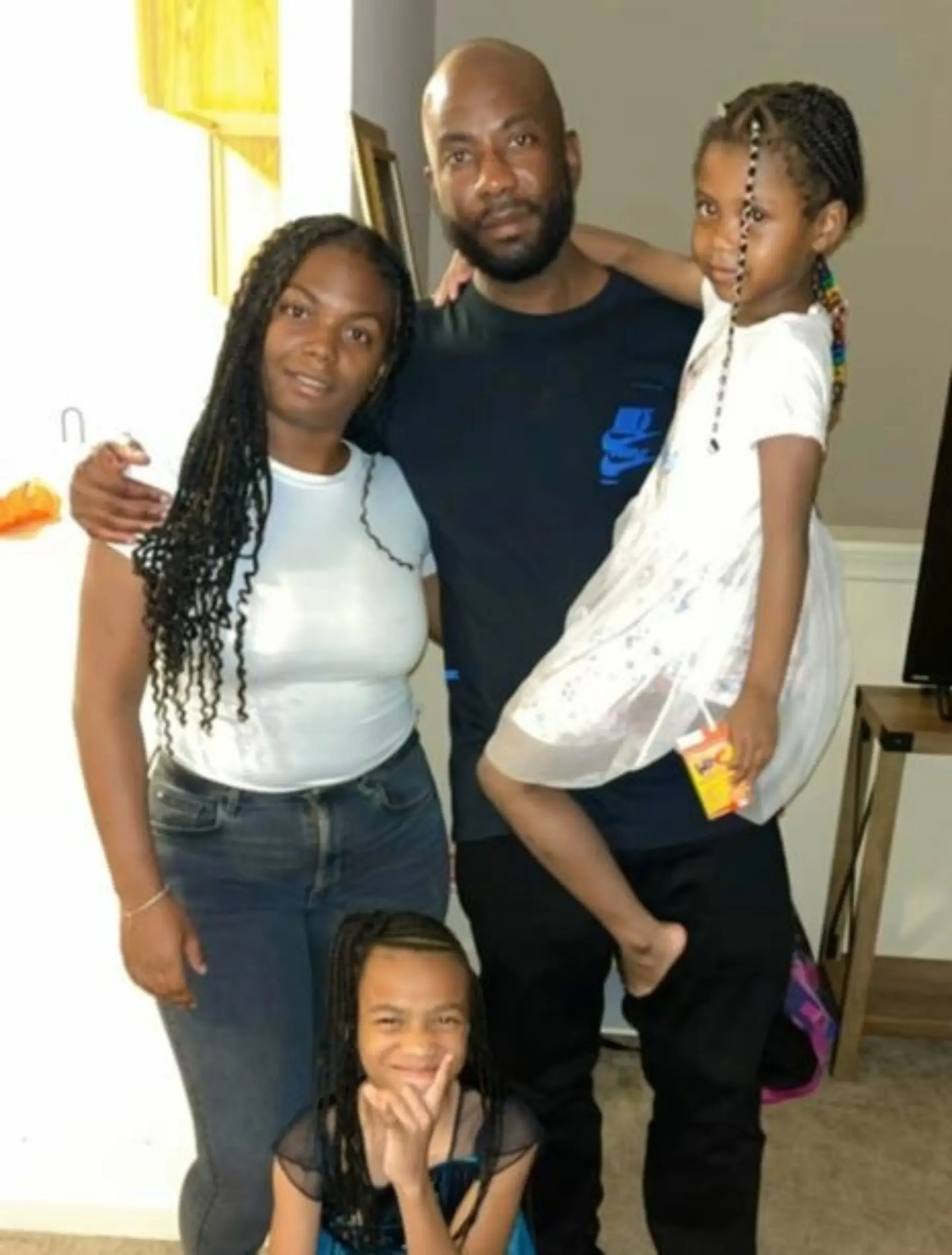 A woman and a man stand together, smiling, with two young girls; one girl is in the man's arms and the other is playfully posing.