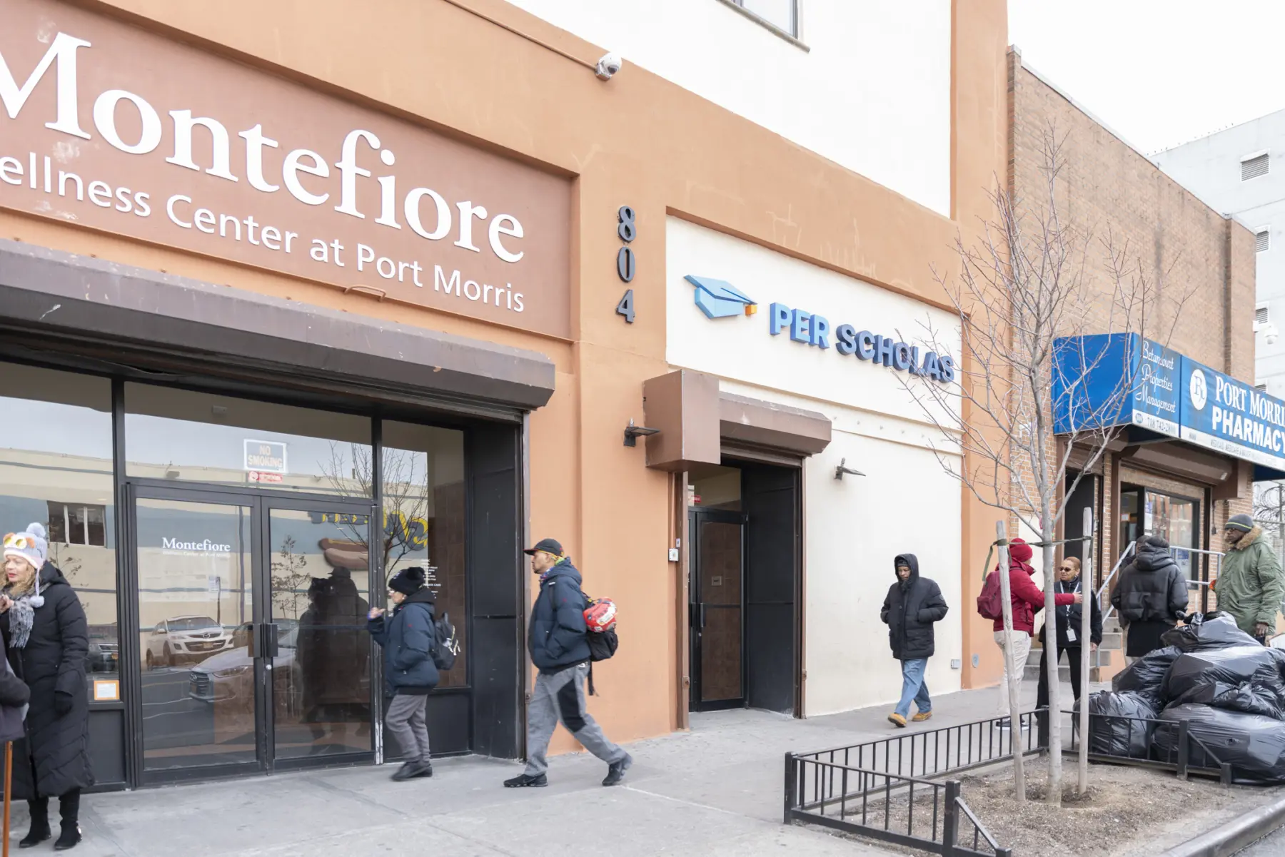 A bustling city street featuring Montefiore Wellness Center and Per Scholas, with pedestrians walking past and a pharmacy nearby.