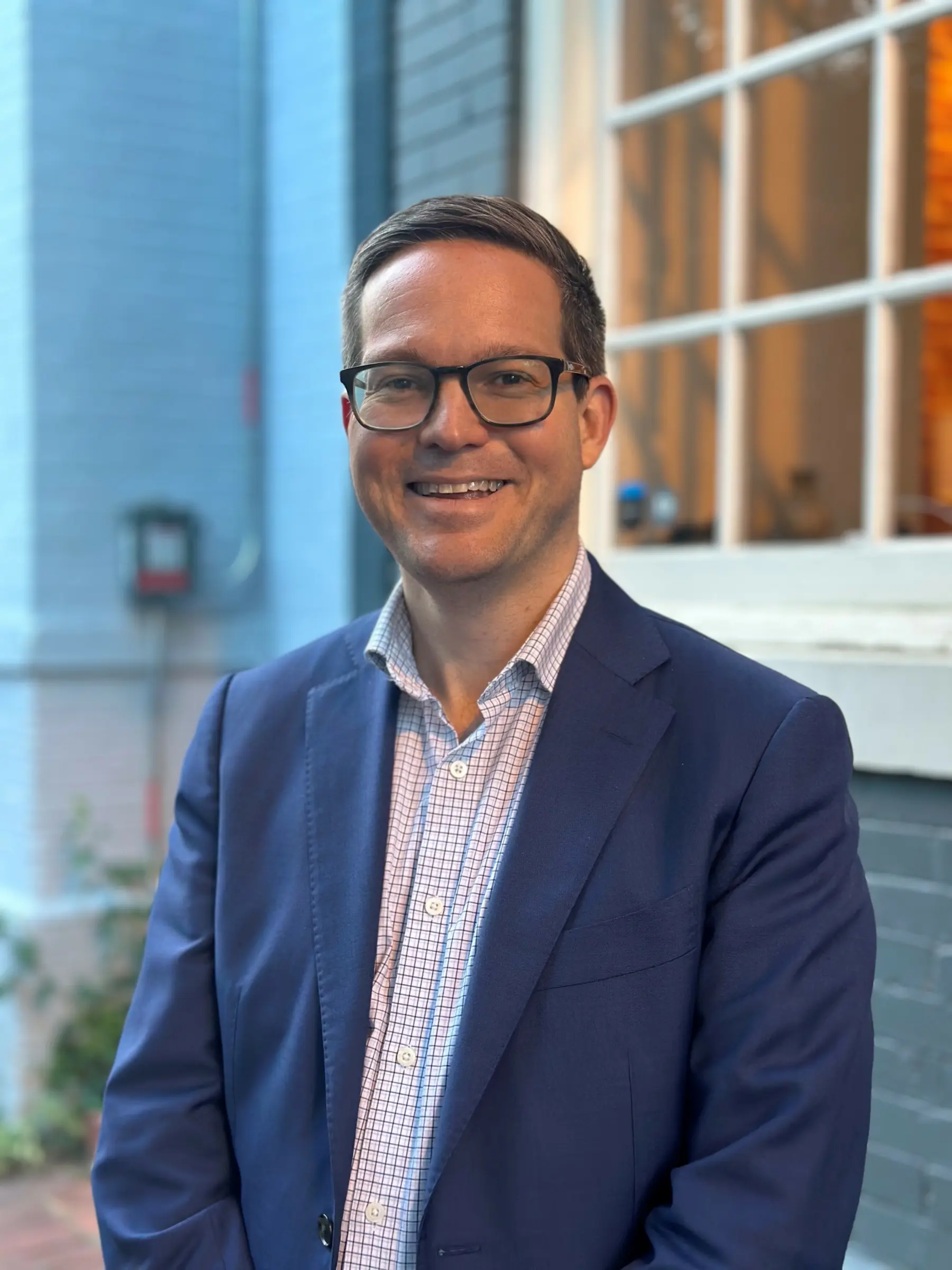 Smiling man in a tailored navy suit stands outside, with a light blue textured wall and a softly lit window in the background.