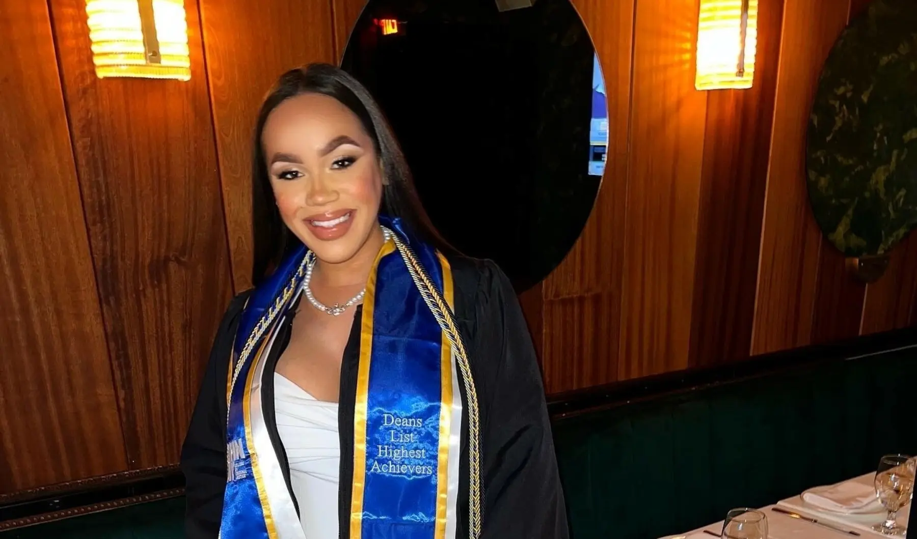 A young woman with long, straight black hair wearing a graduation gown and a blue banner around her neck stating she was on the dean's list. She is standing in a restaurant with two lights and a mirror behind her.