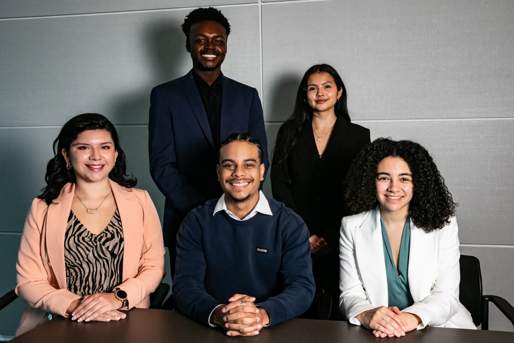 A diverse group of five young adults, dressed in professional attire, poses together, smiling confidently in a modern office setting.