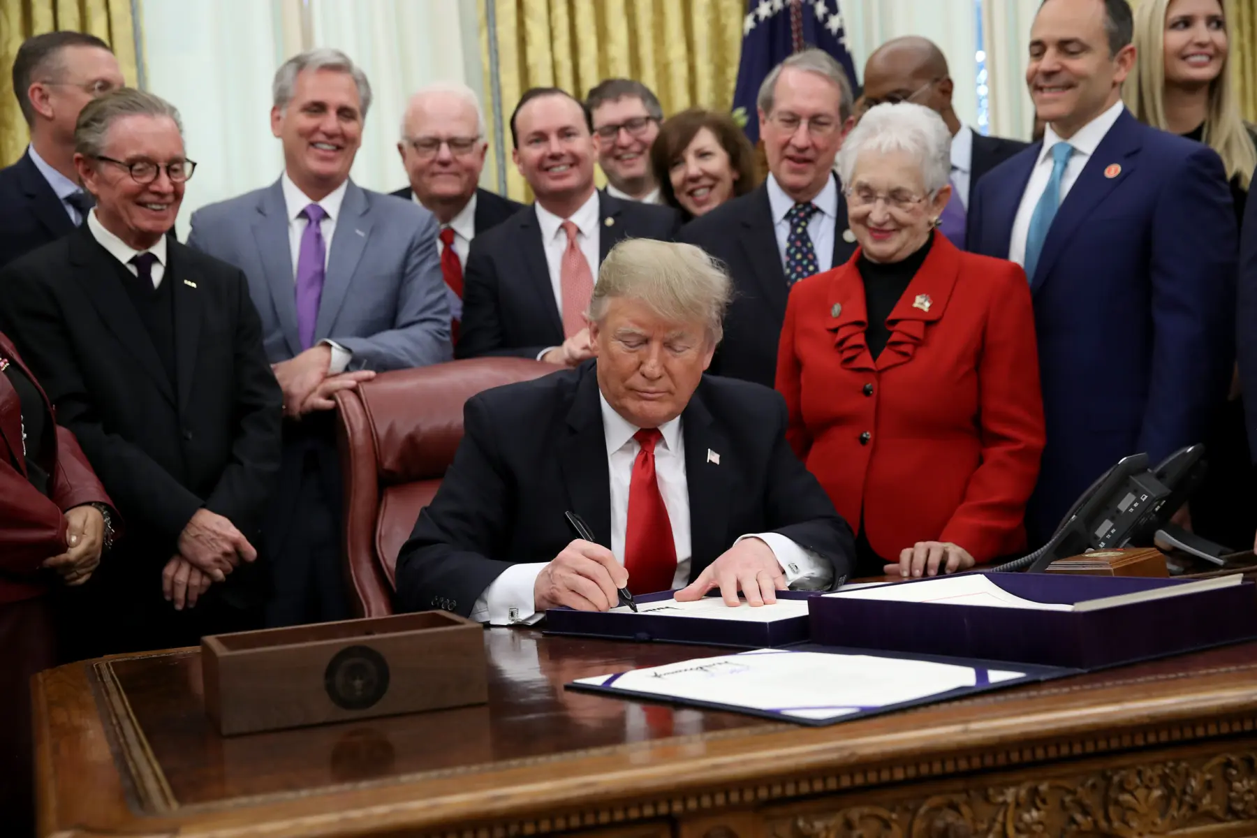 Donald Trump signs a document at a desk, surrounded by smiling officials in suits, capturing a moment of legislative celebration.