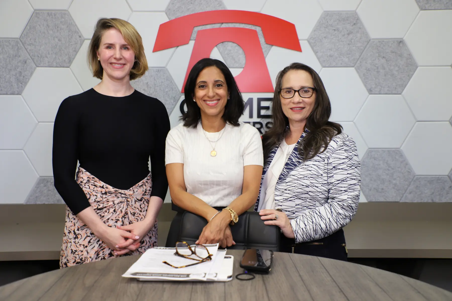 Three women stand together, smiling, in a modern office setting with a hexagonal wall design. Papers and glasses are on the table.