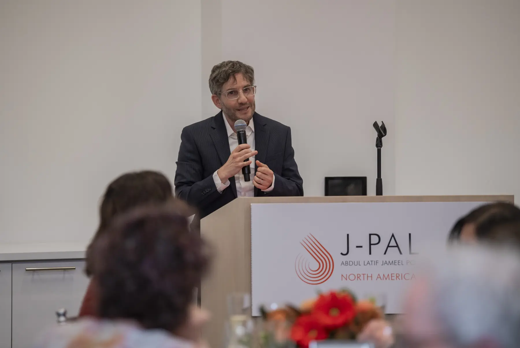 A speaker in a suit addresses an audience from behind a podium with the J-PAL logo, promoting engagement at a professional event.
