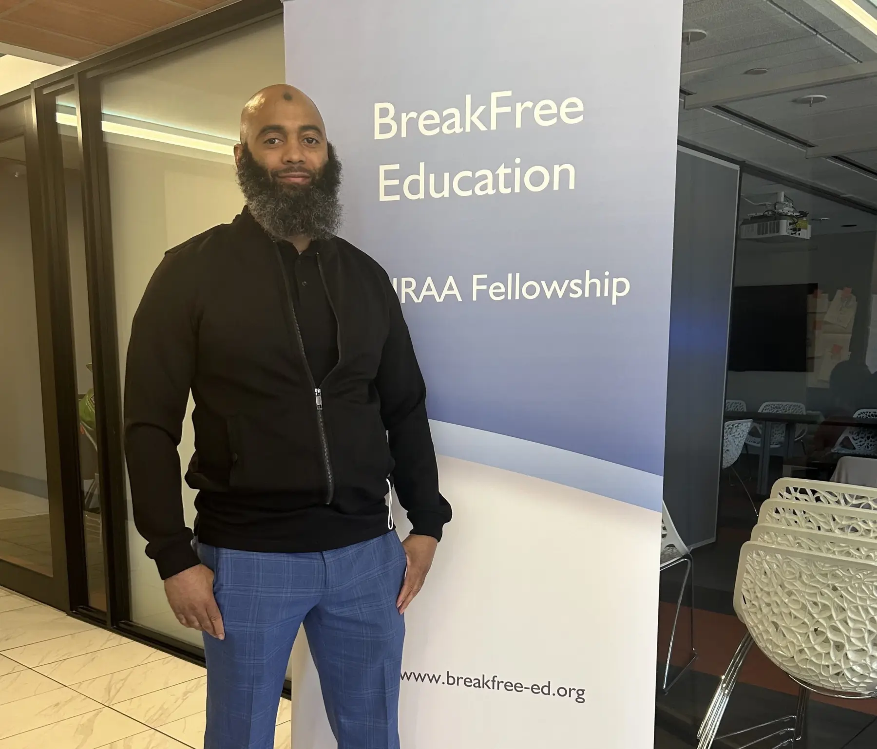 Image of a tall Black man with a bald head and a full beard wearing a black sweater and blue pants, standing in front of a banner that says "BreakFree Education"