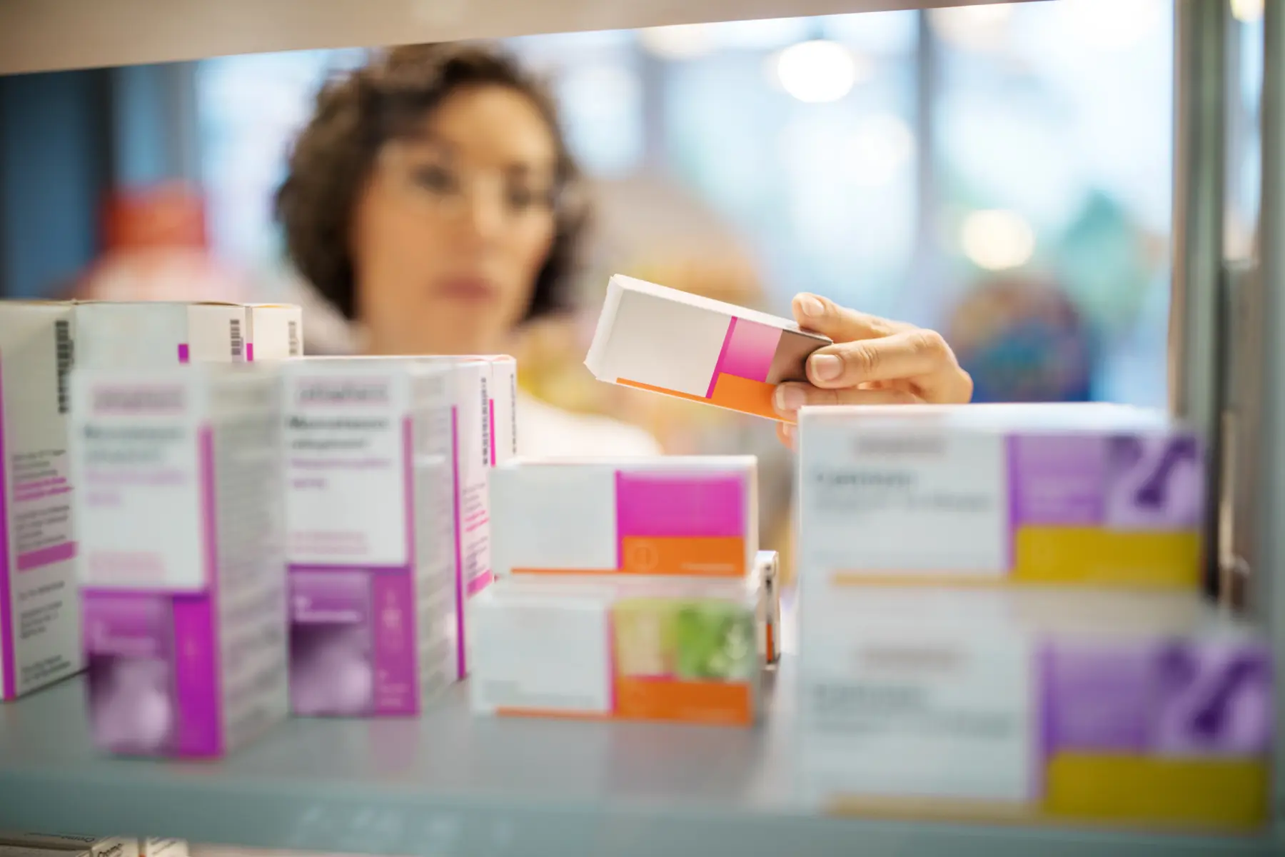 A pharmacist examines a medication box with pink accents while shelves filled with various other pharmaceutical products are visible.