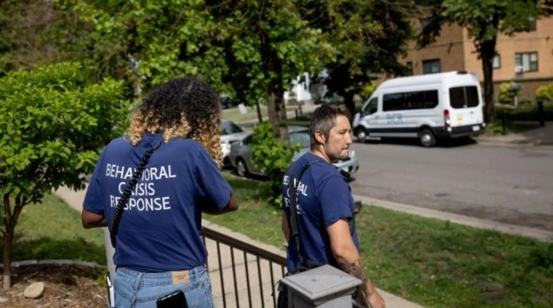 Two members of a behavioral crisis response team, wearing navy shirts, walk down steps towards a residential street with trees.