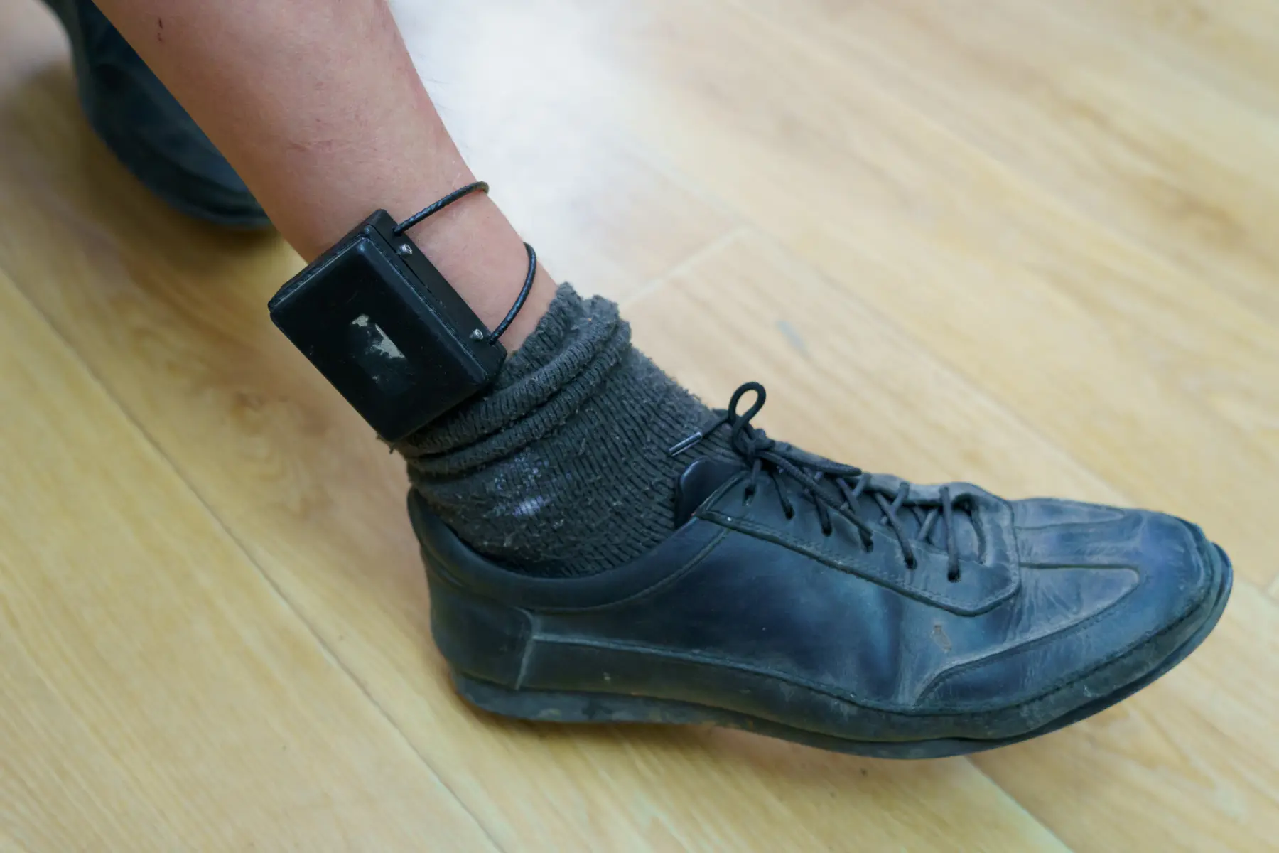 A black electronic monitoring device is attached to a person's ankle, alongside a black shoe and dark sock on a wooden floor.