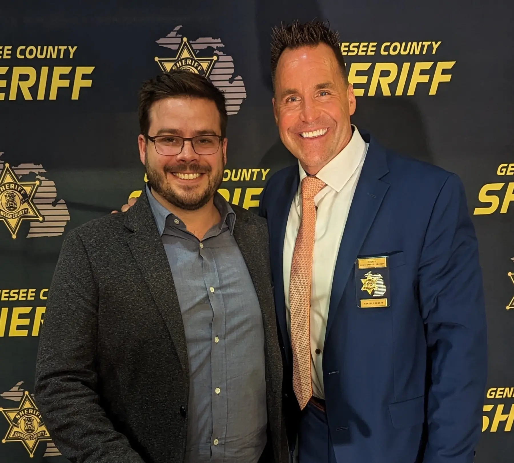 Two men pose together, one in a gray blazer and glasses, the other in a blue suit with an orange tie, smiling against a sheriff's backdrop.