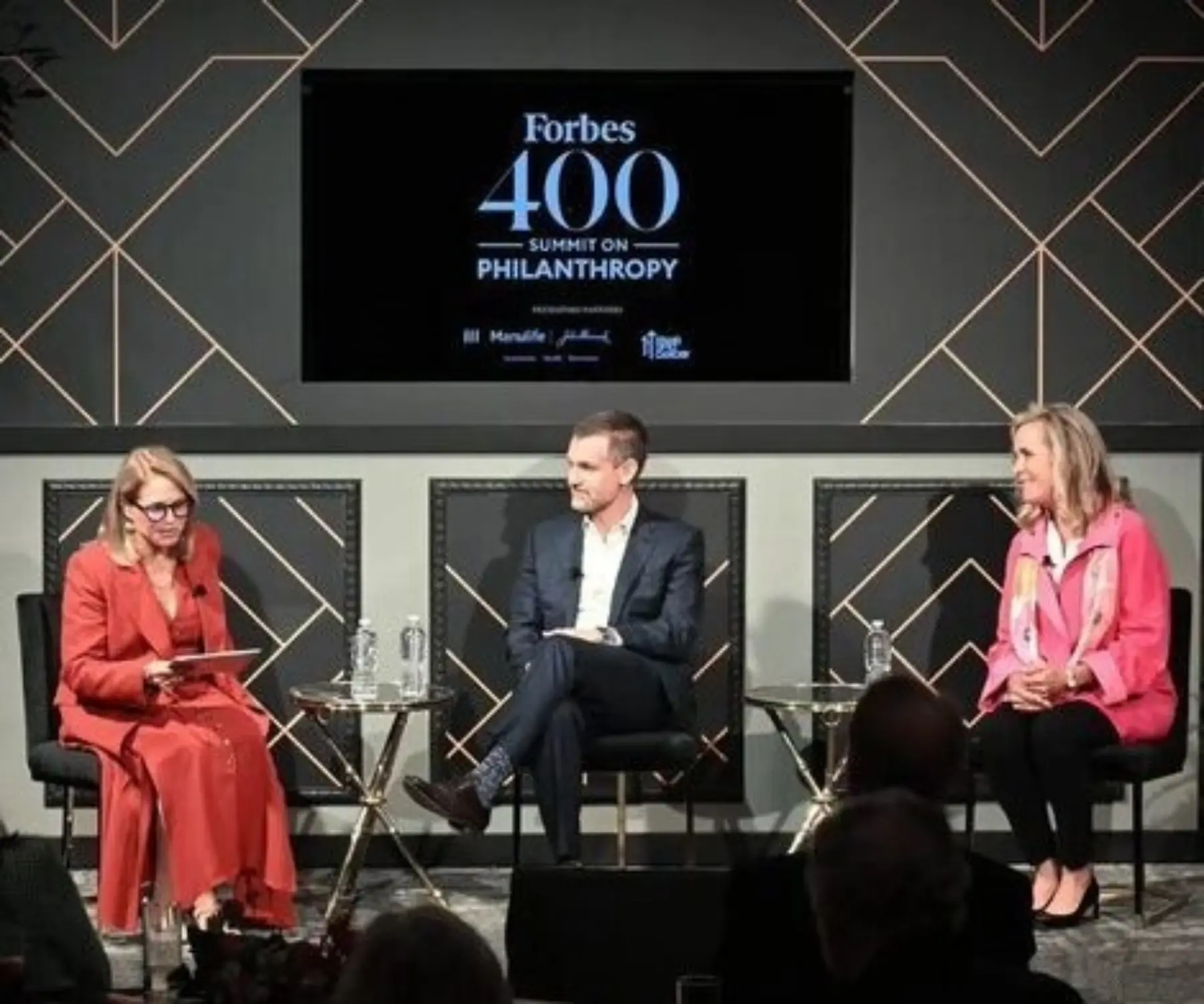 A panel discussion featuring three speakers: a woman in a red dress, a man in a suit, and a woman in a pink scarf, discussing philanthropy.