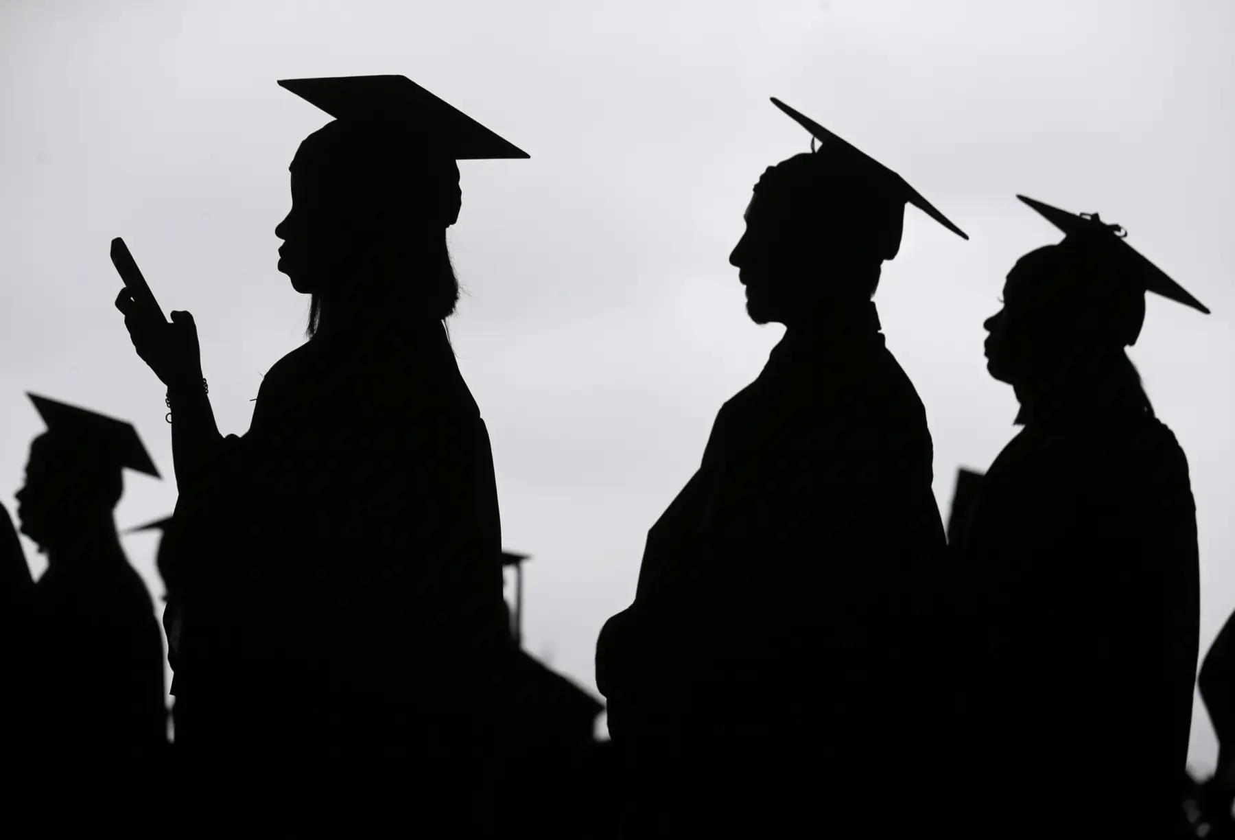 Silhouettes of graduates wearing caps stand in profile, one checking a phone, against a cloudy sky, capturing a moment of anticipation.