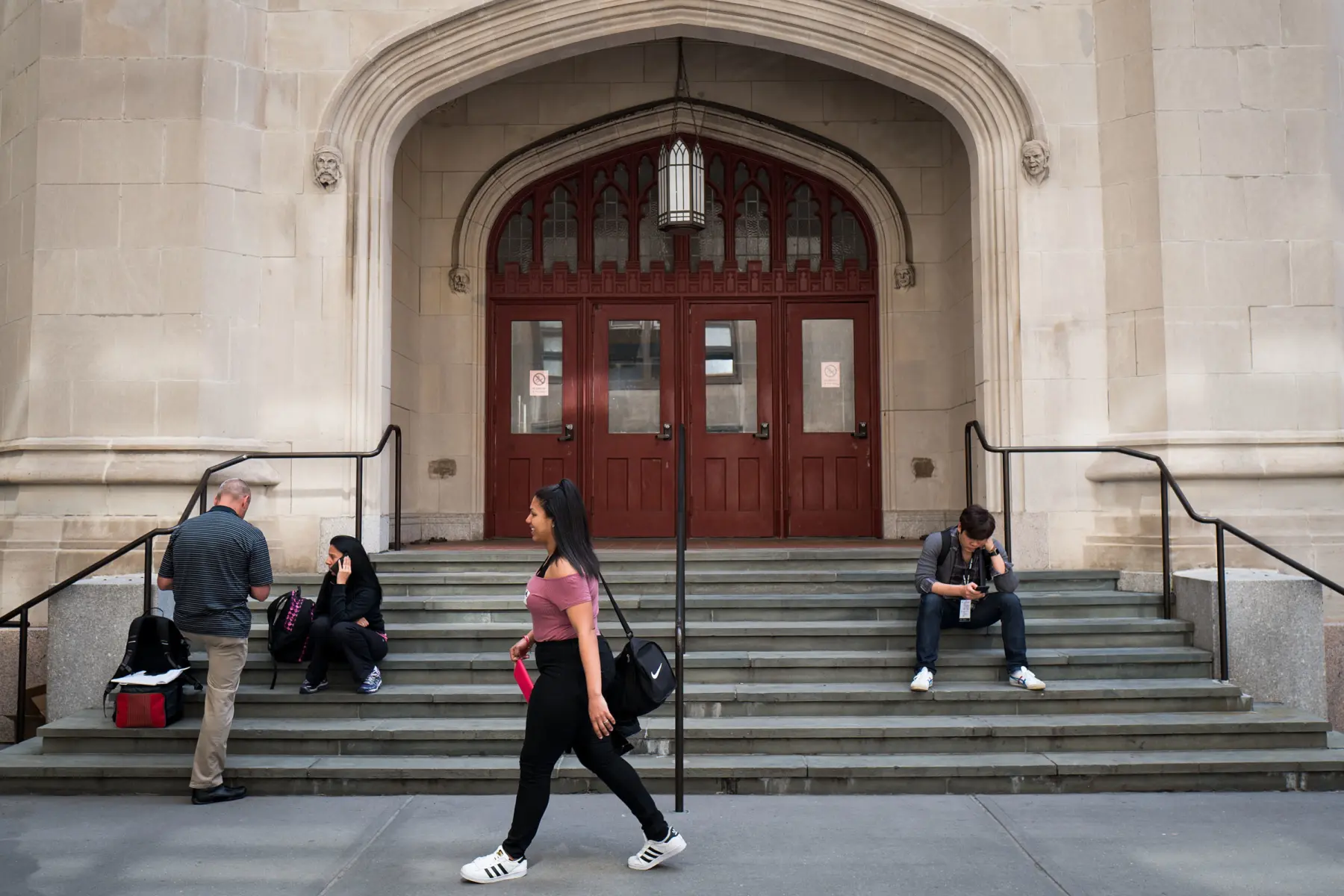 A group of students gathers on stone steps outside a building, one walking by while others sit and engage with their phones.
