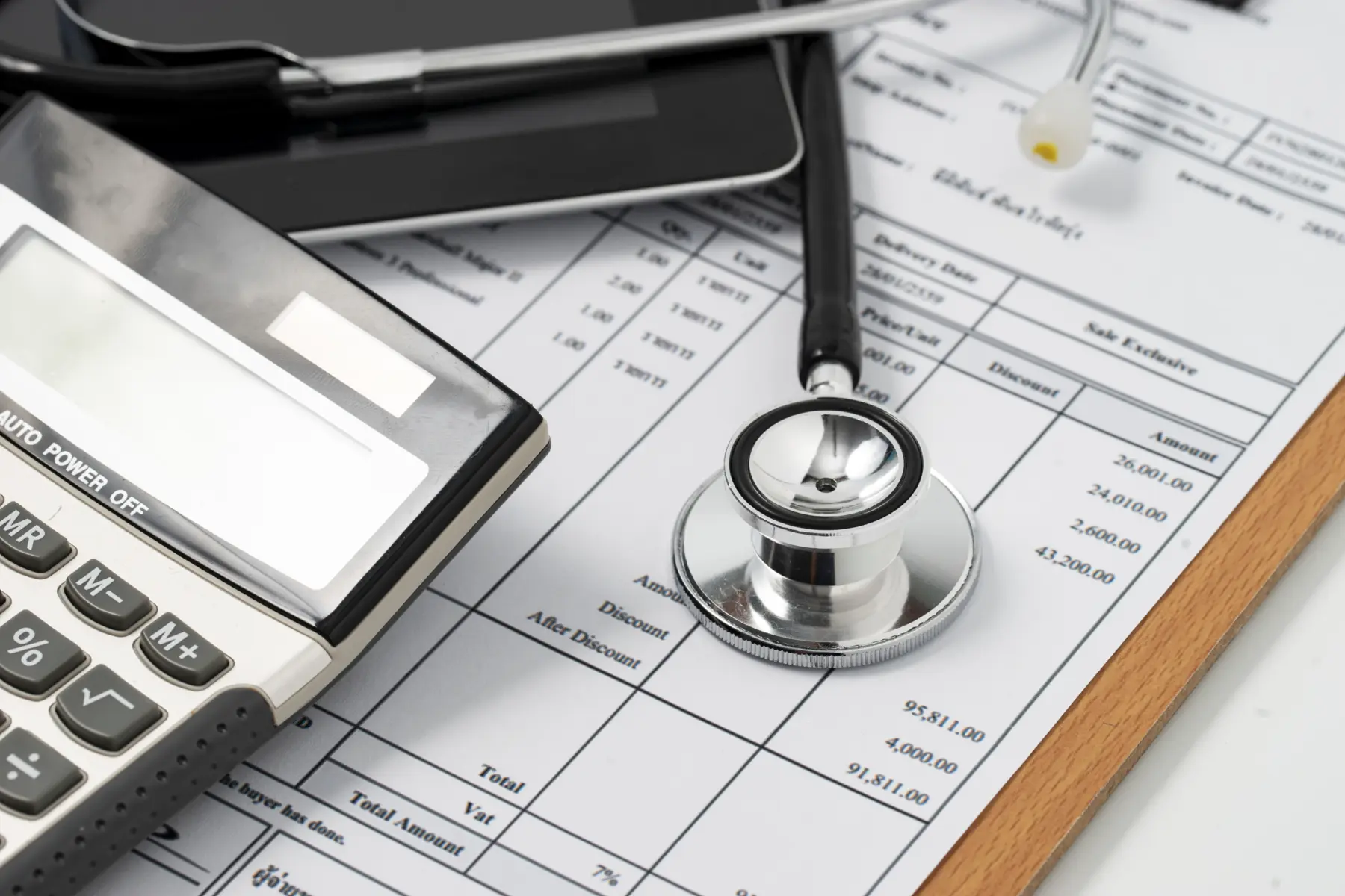 A calculator, stethoscope, and tablet rest atop a detailed invoice form with financial figures and columns clearly visible.