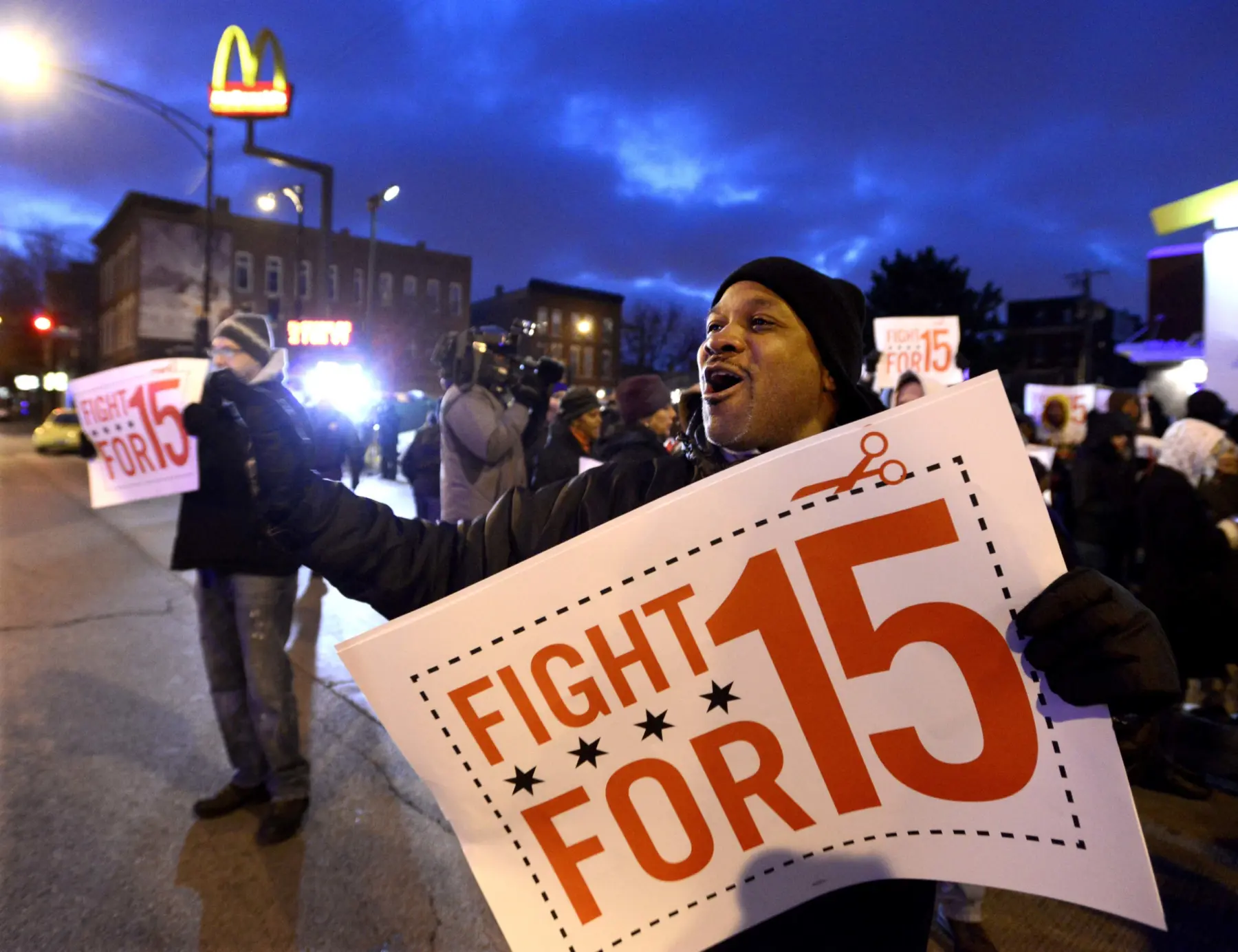 A crowd rallies at dusk, holding signs that read "Fight for 15," advocating for higher wages, with a McDonald's sign in the background.