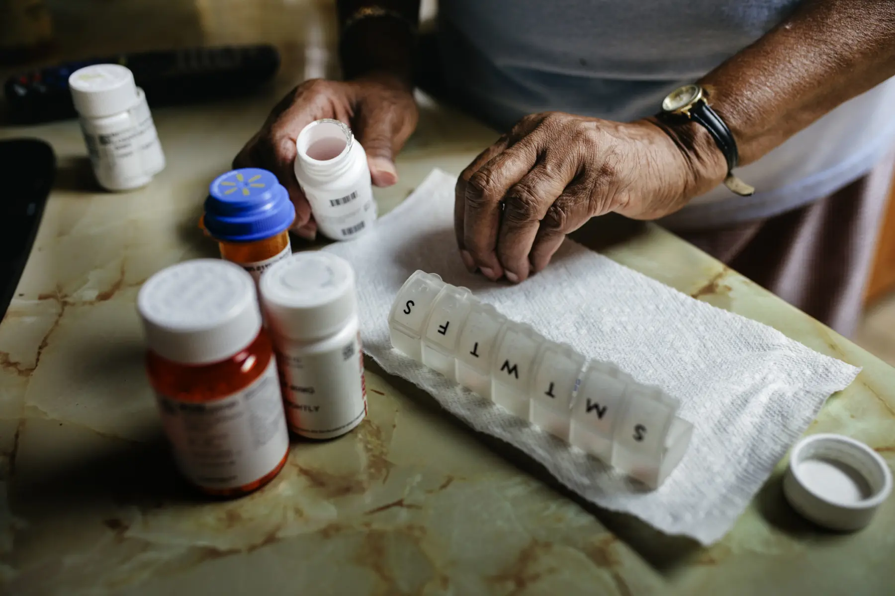 Hands sort through various prescription bottles on a countertop, arranging pills into a weekly organizer for medication management.