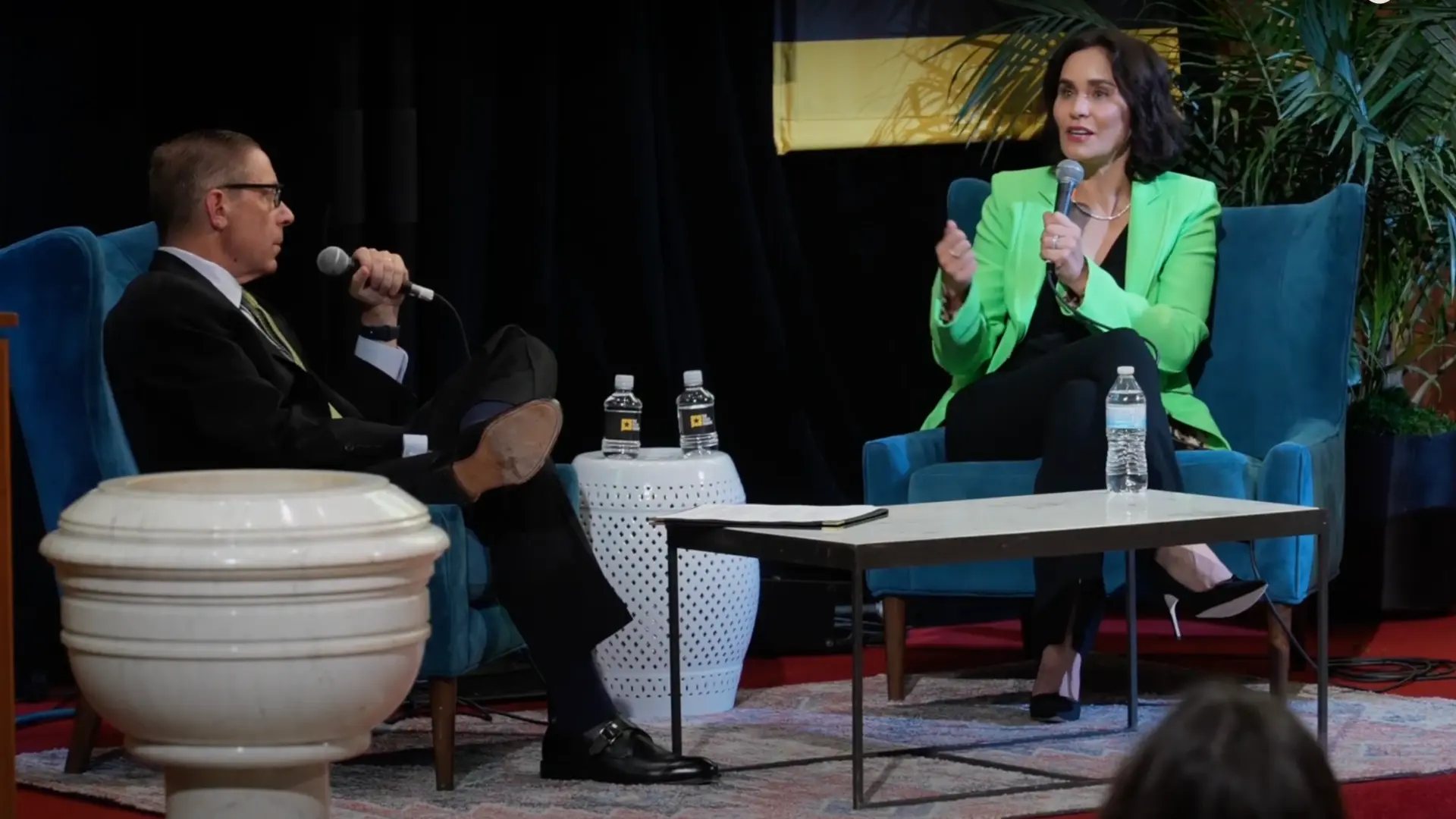 A man and woman engage in a dialogue while seated in stylish chairs, with a small table displaying water bottles and a notepad between them.