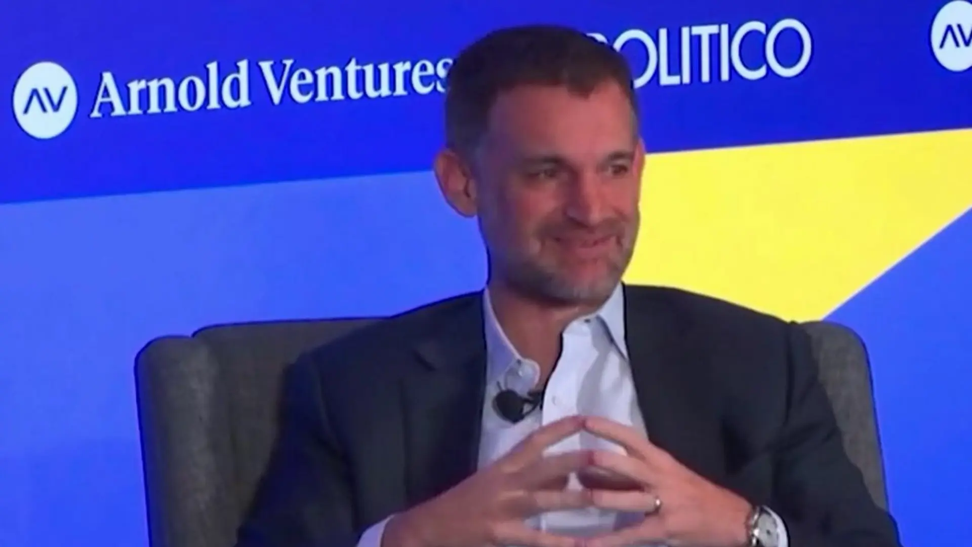A man in a suit sits thoughtfully, hands clasped, against a backdrop featuring the logos of Arnold Ventures and Politico.